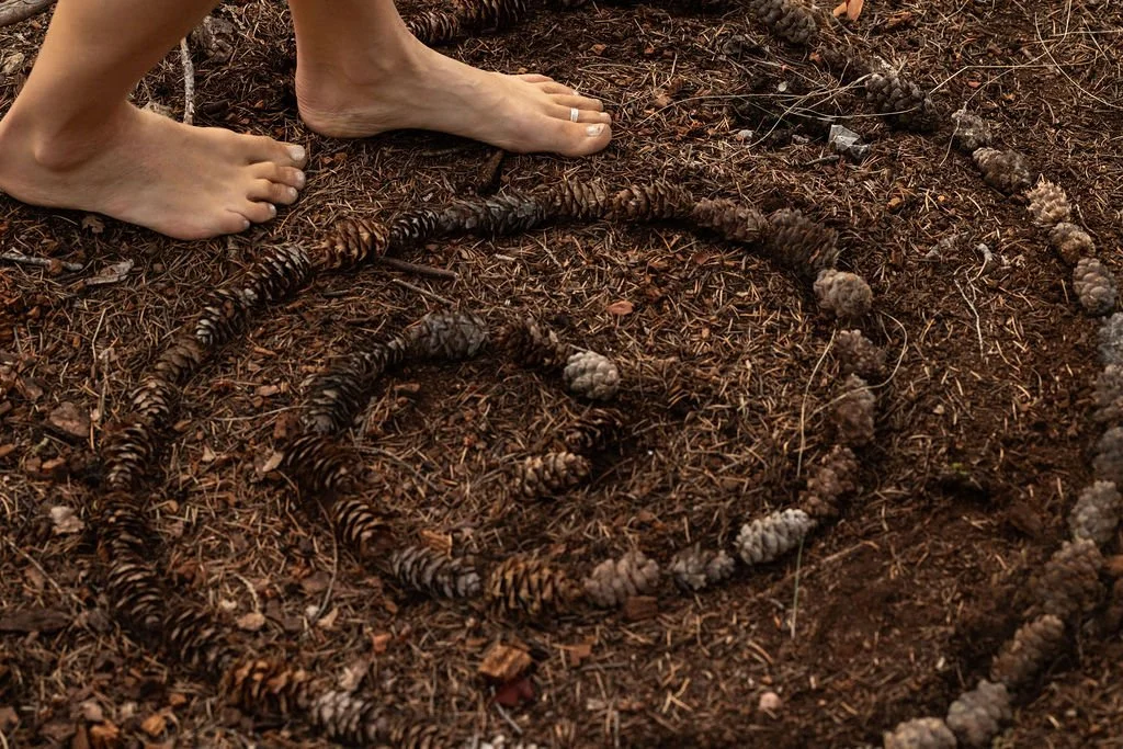 A person's bare feet standing next to a spiral pattern made of pinecones on the forest floor.