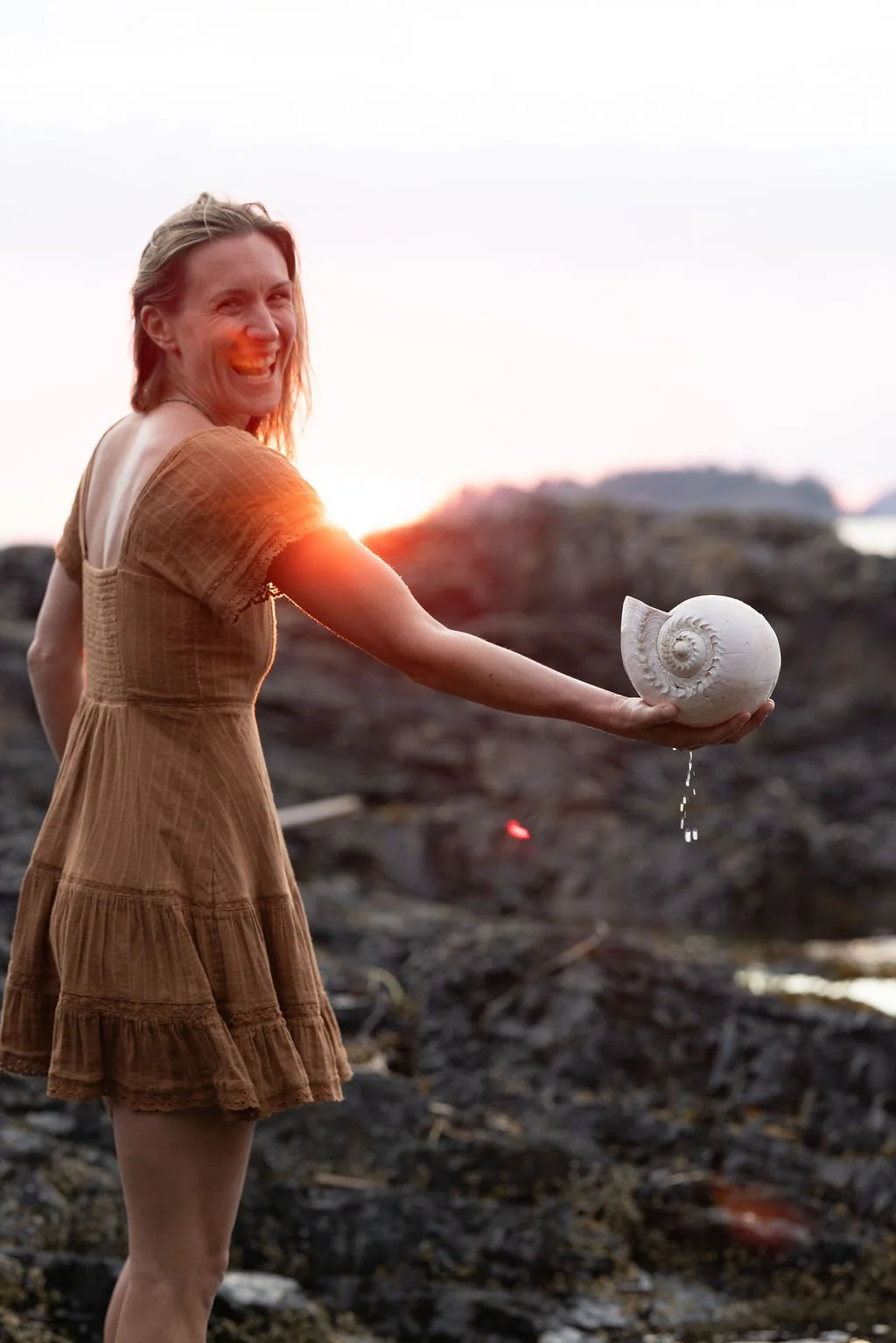 A woman smiling while holding a spiraled shell with water dripping from it, outdoors during sunset.
