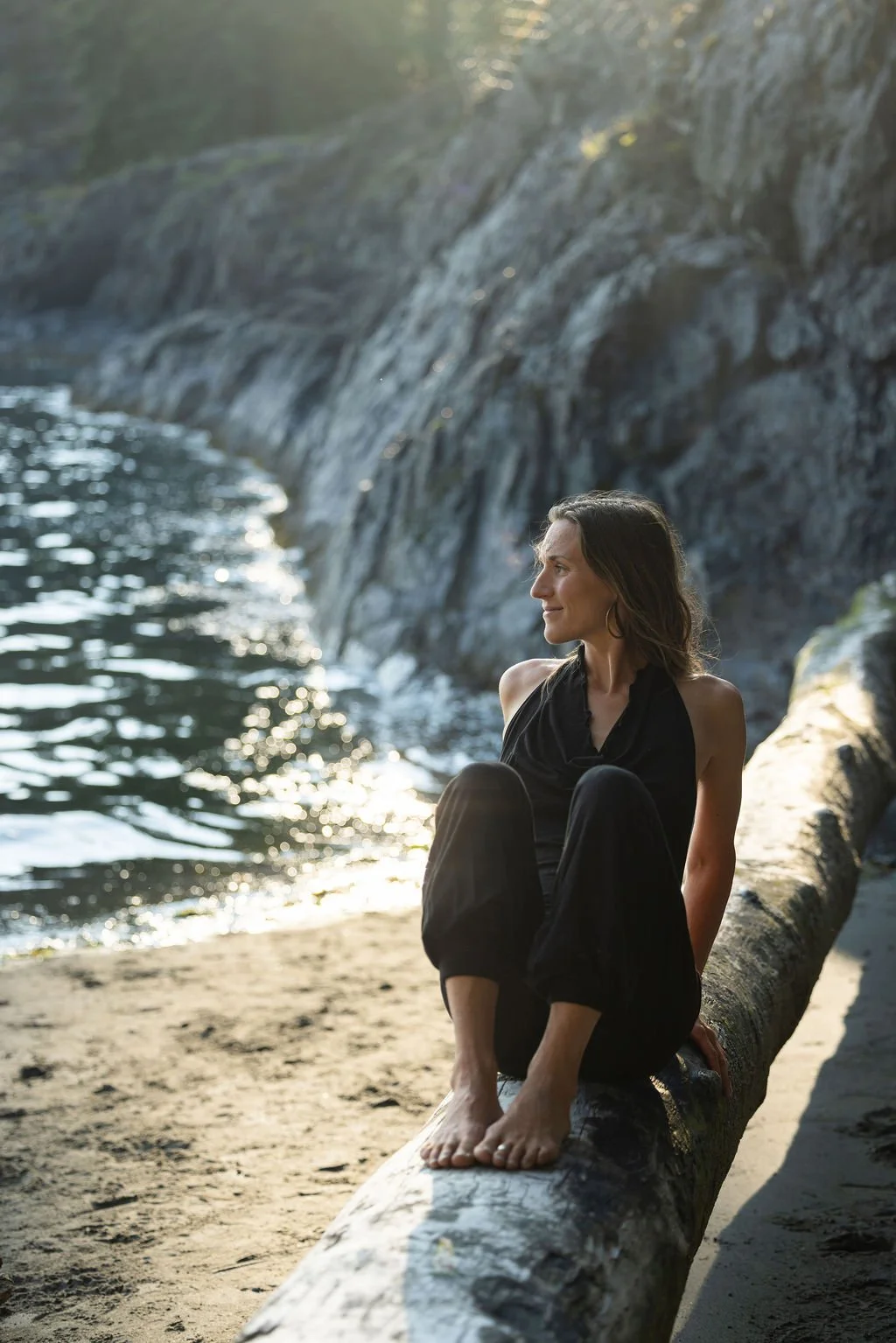 A woman in black clothing sitting barefoot on a large fallen tree trunk on a beach, with rocky cliffs and water in the background, illuminated by sunlight.