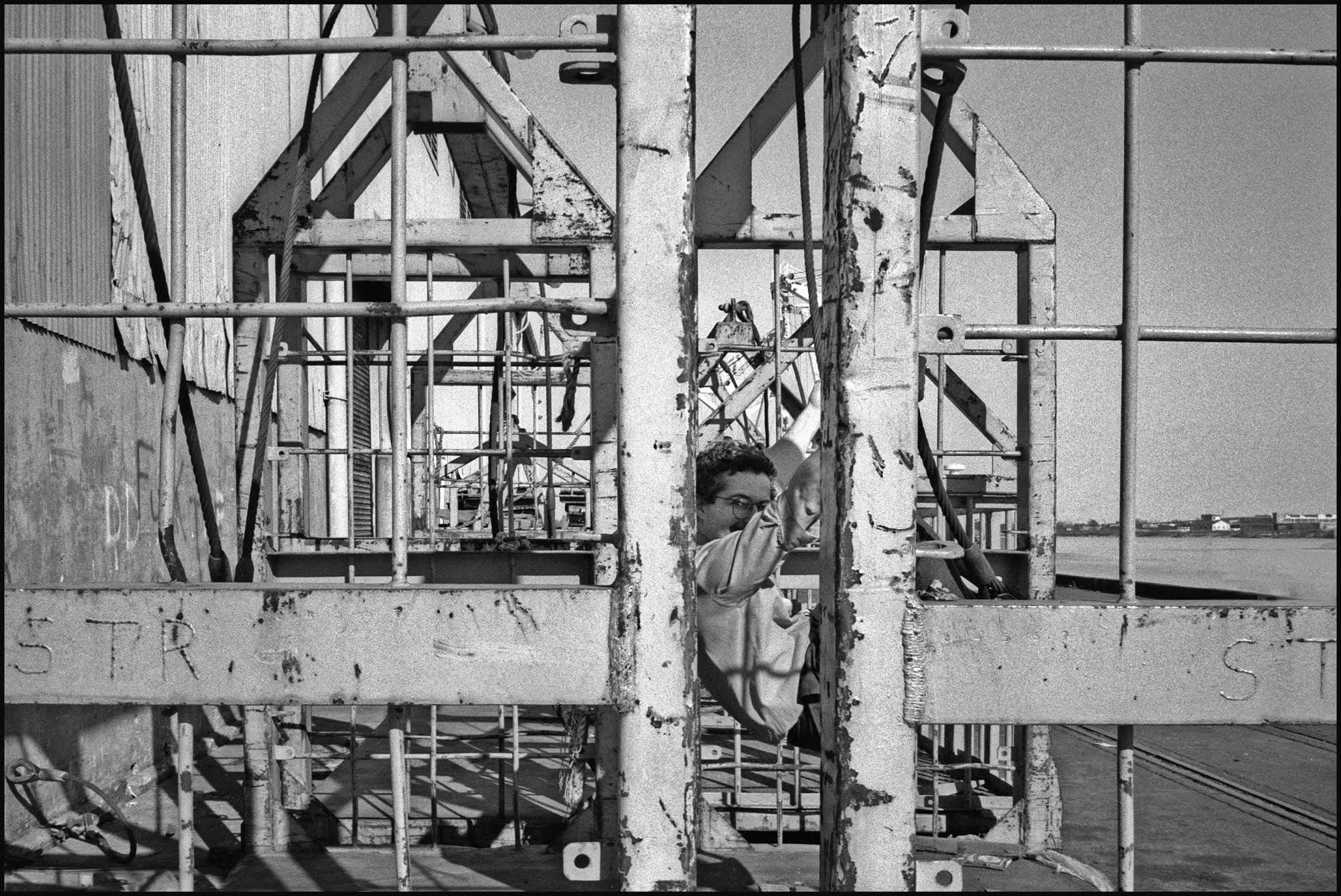 Black and white photo of Douglas Darden crouching in metal scaffolding near a body of water, with a clear sky.