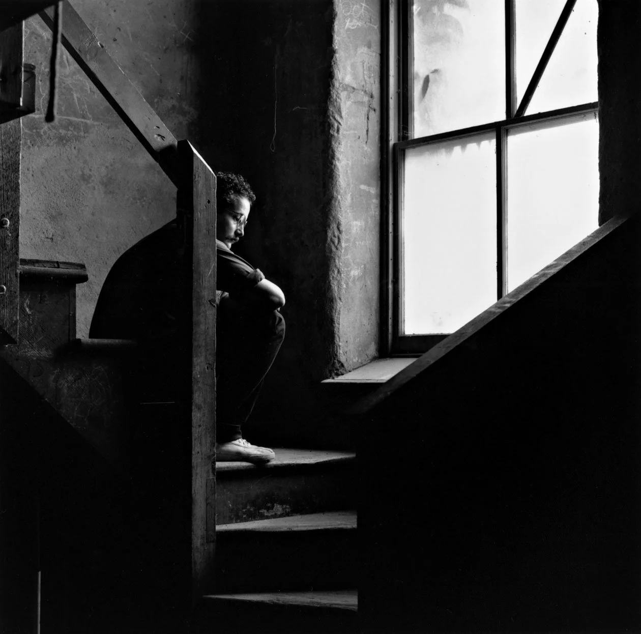 Douglas Darden sitting on a staircase, looking down with a contemplative expression, near a large window with frosted glass, in black and white.
