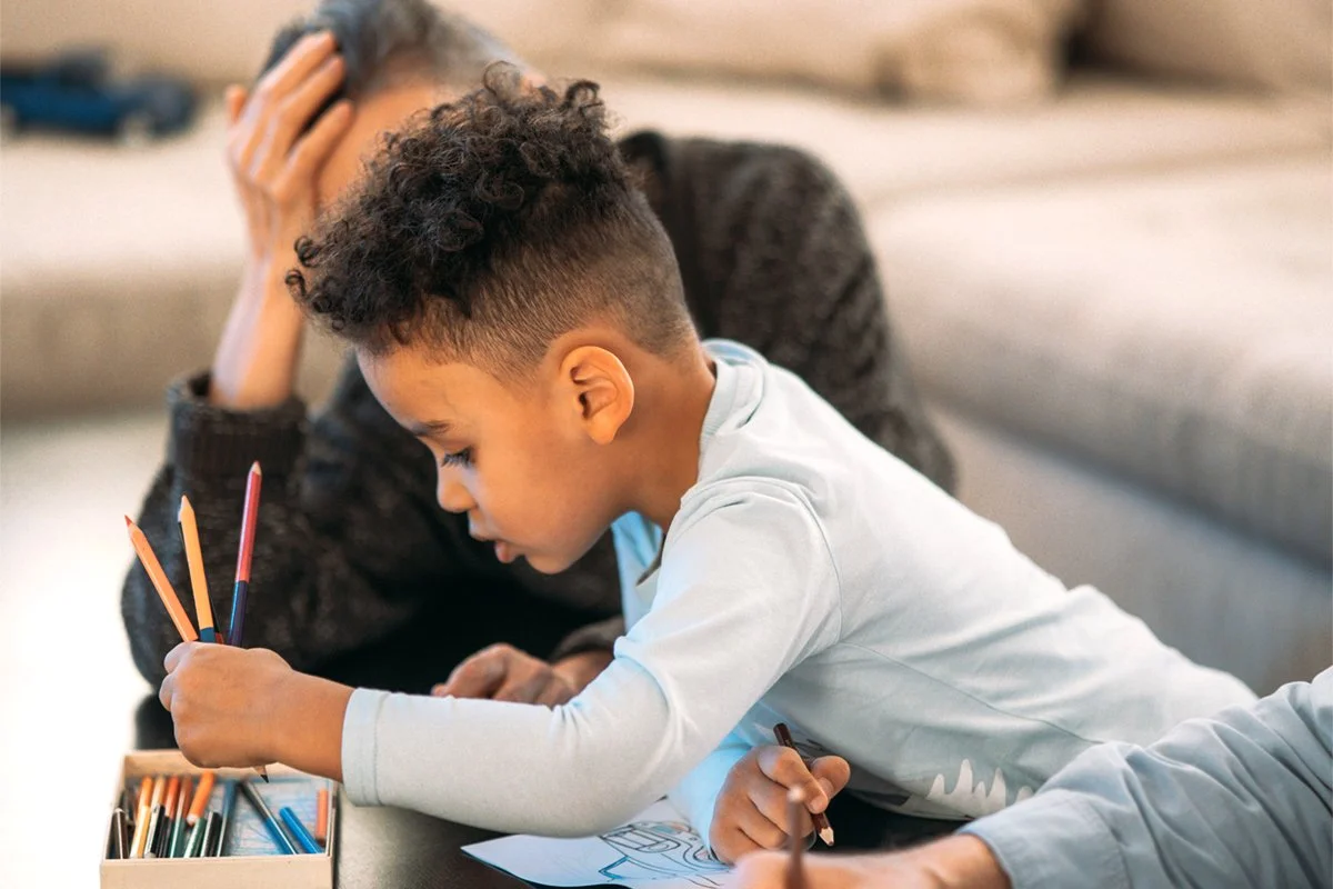 A young boy coloring with colored pencils at a table and a counselor seated nearby helping and supervising him.