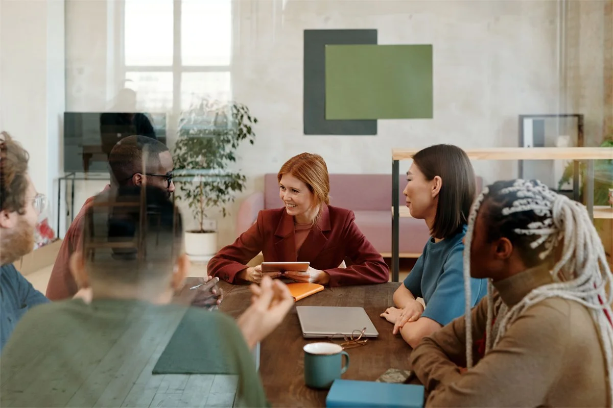 A diverse group of six people sitting around a table in a modern office, engaged in a discussion, with a woman in a red blazer speaking and others listening.