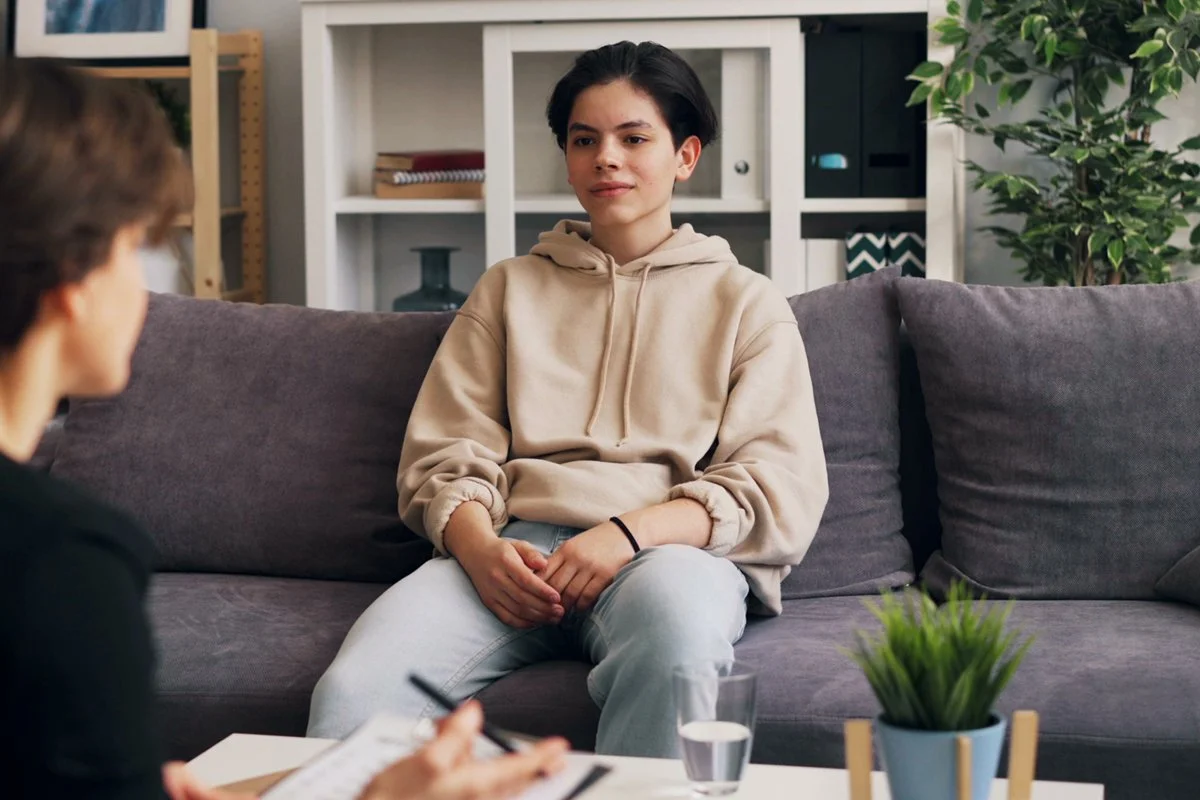 A teenager hair sitting on a gray sofa during a therapy or counseling session, with a therapist and a notepad visible in the foreground.