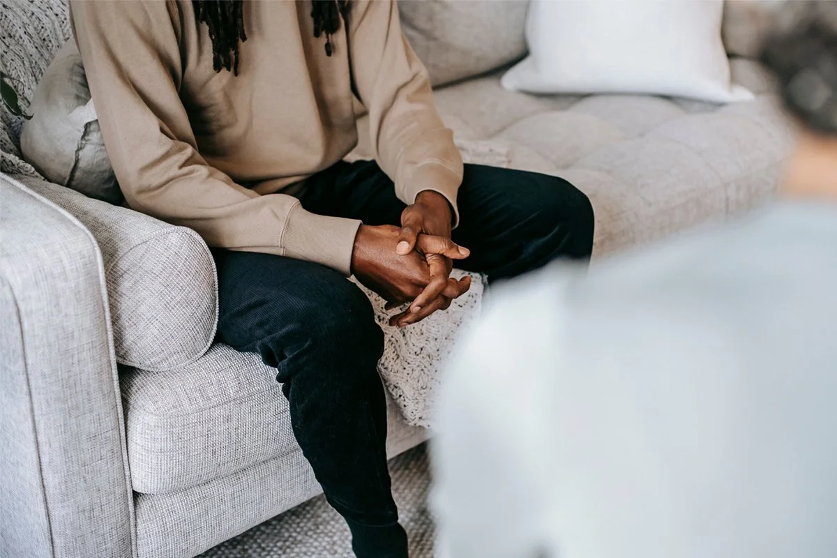 A person sitting on a light-colored fabric sofa with their hands clasped together in their lap, engaged in conversation with someone whose slightly visible in the foreground.