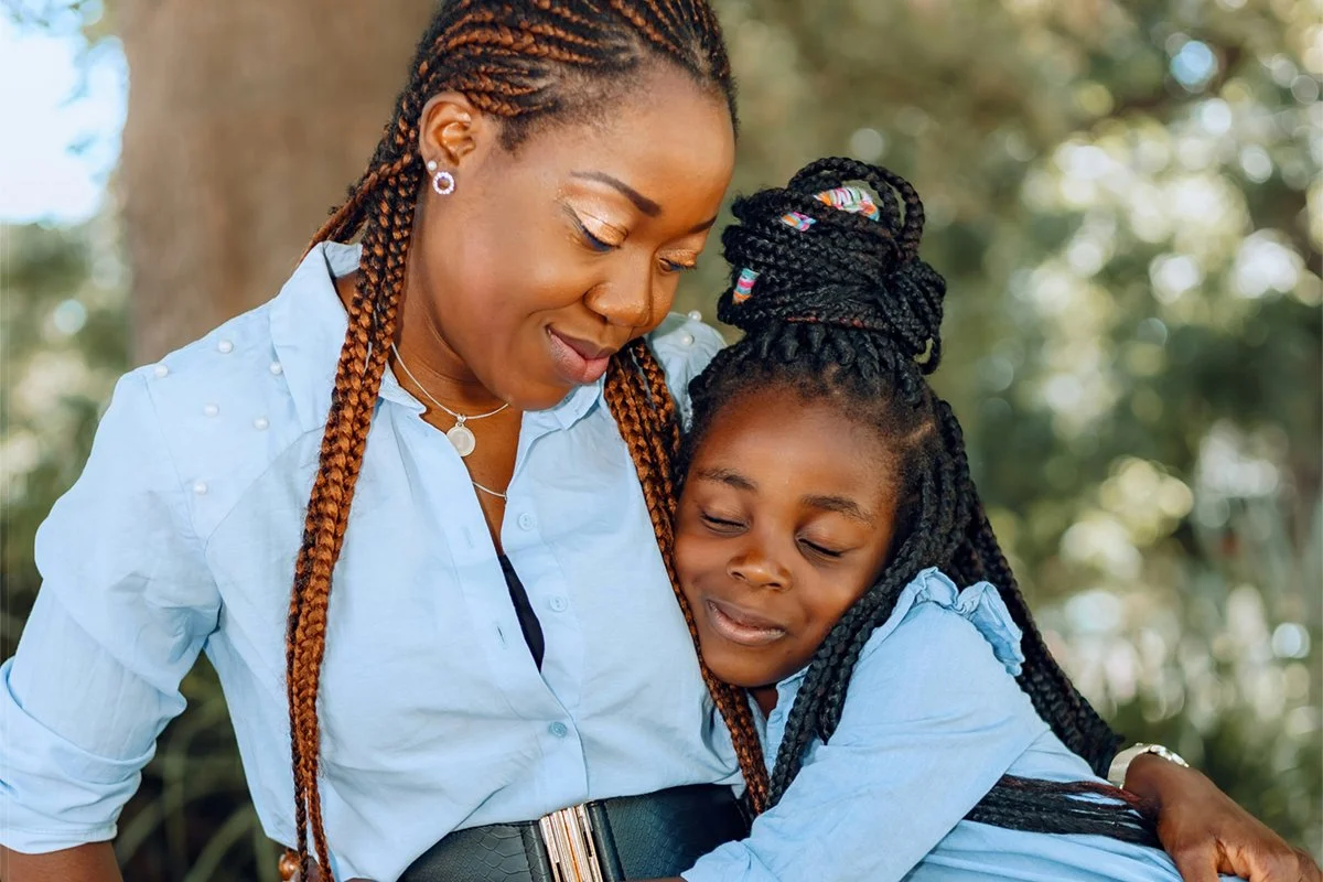 A mother and child hugging outdoors, both with closed eyes and smiling gently.