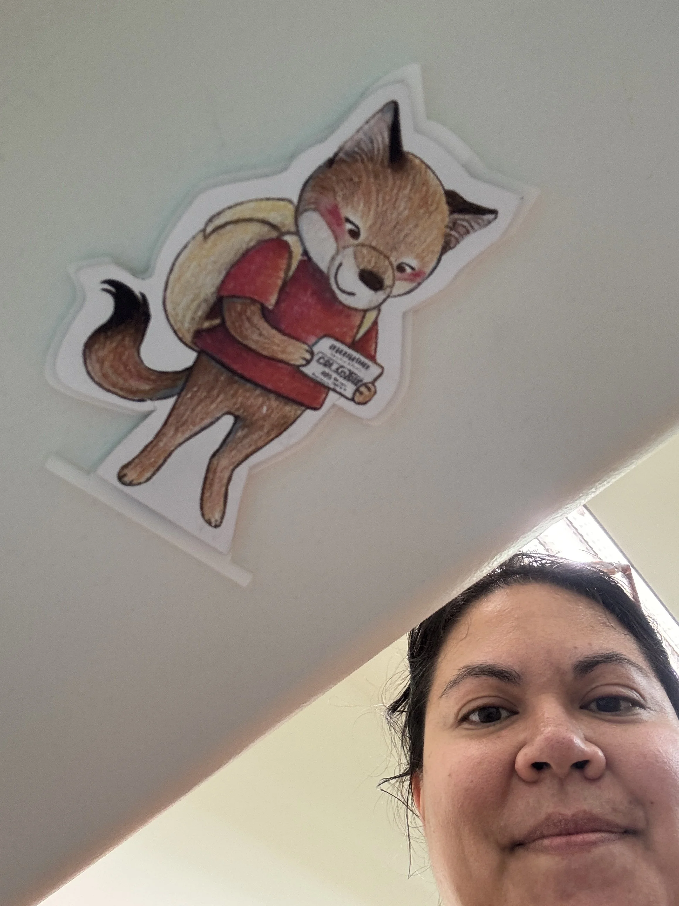A woman taking a selfie with her face partially visible in the lower right corner of the image. There is a paper cutout of Odi the Coyote with a backpack and holding a library card hanging on the bottom side of a window sill.