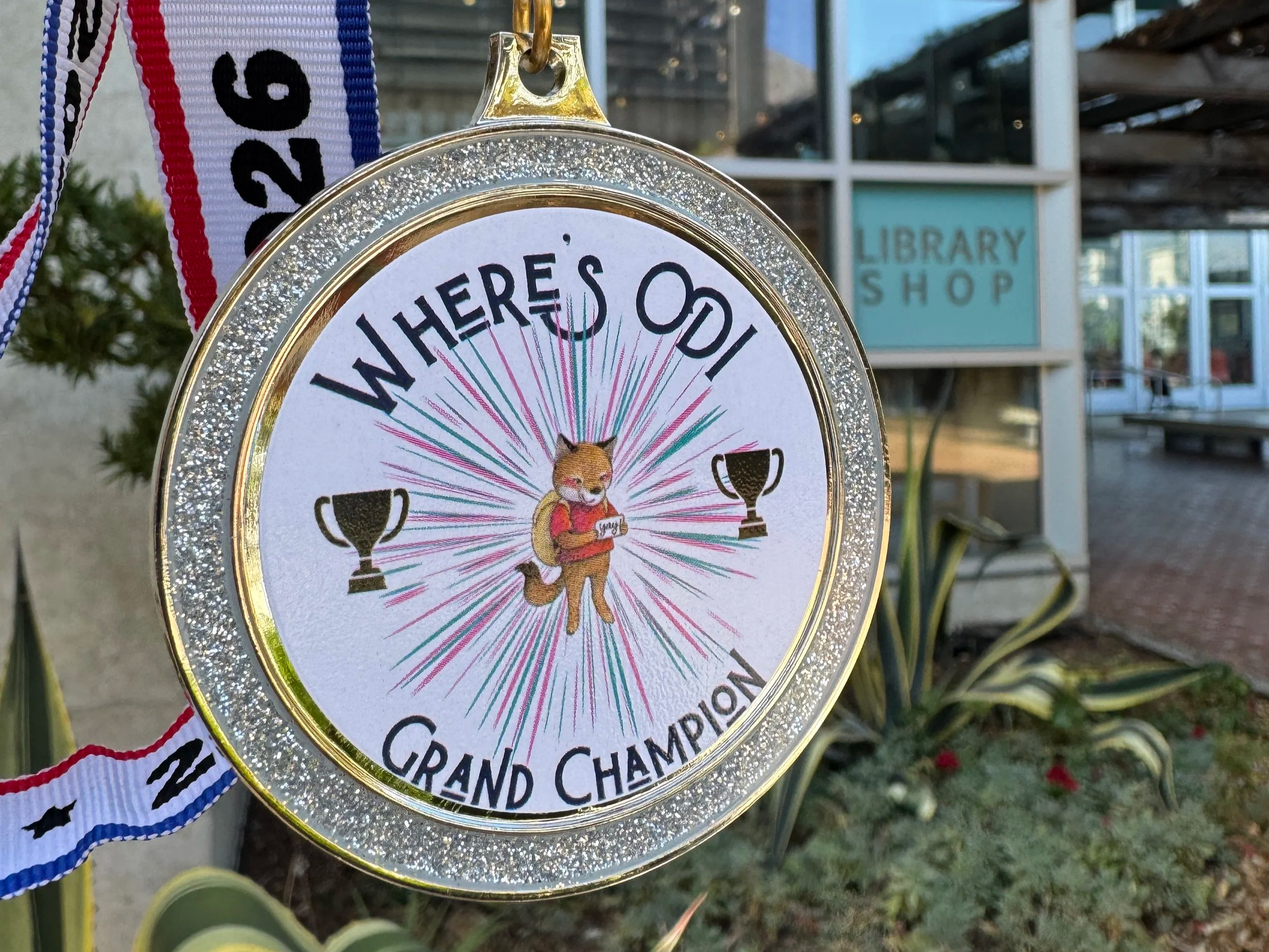 Gold and glittery medal with a ribbon displaying the year 2026, awarded for a grand champion event, the Where's Odi challenge, hung outdoors in front of the Library Shop where it was obtained.