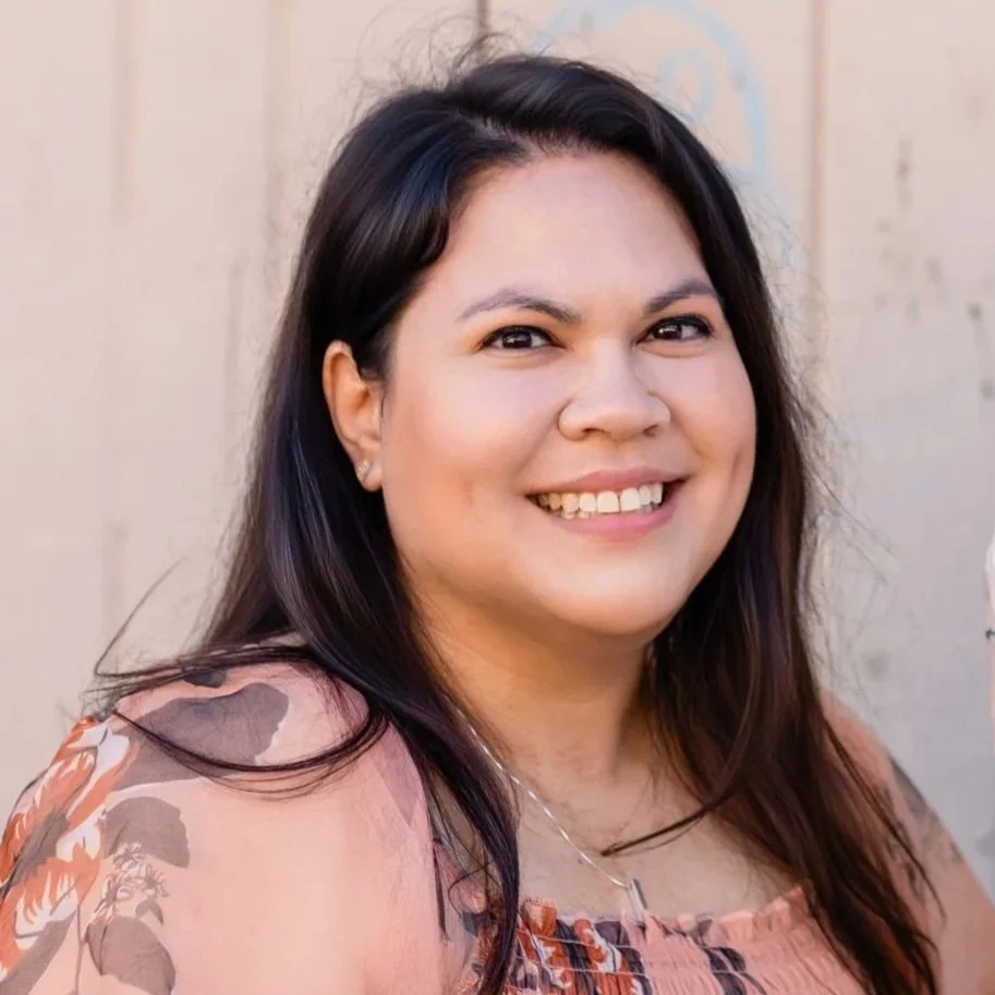 This is Jen, the author of this website. In this photo, she is pictures smiling. She has dark hair and is wearing a patterned blouse. The photo was taken outdoors in front of a weathered wall.