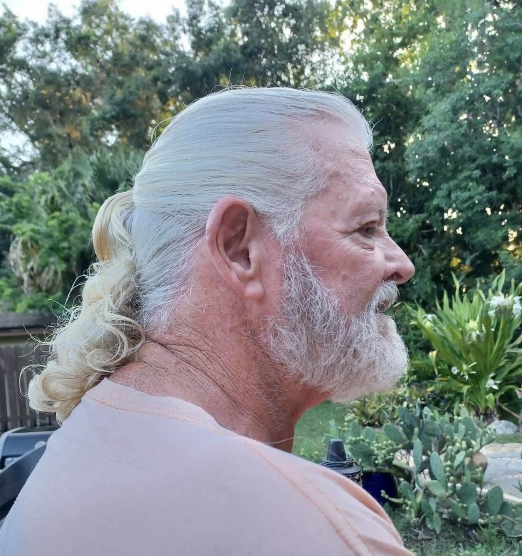 Close-up side view of an elderly man's face with long white hair and beard, wearing a light-colored shirt, with a garden background of greenery and trees.