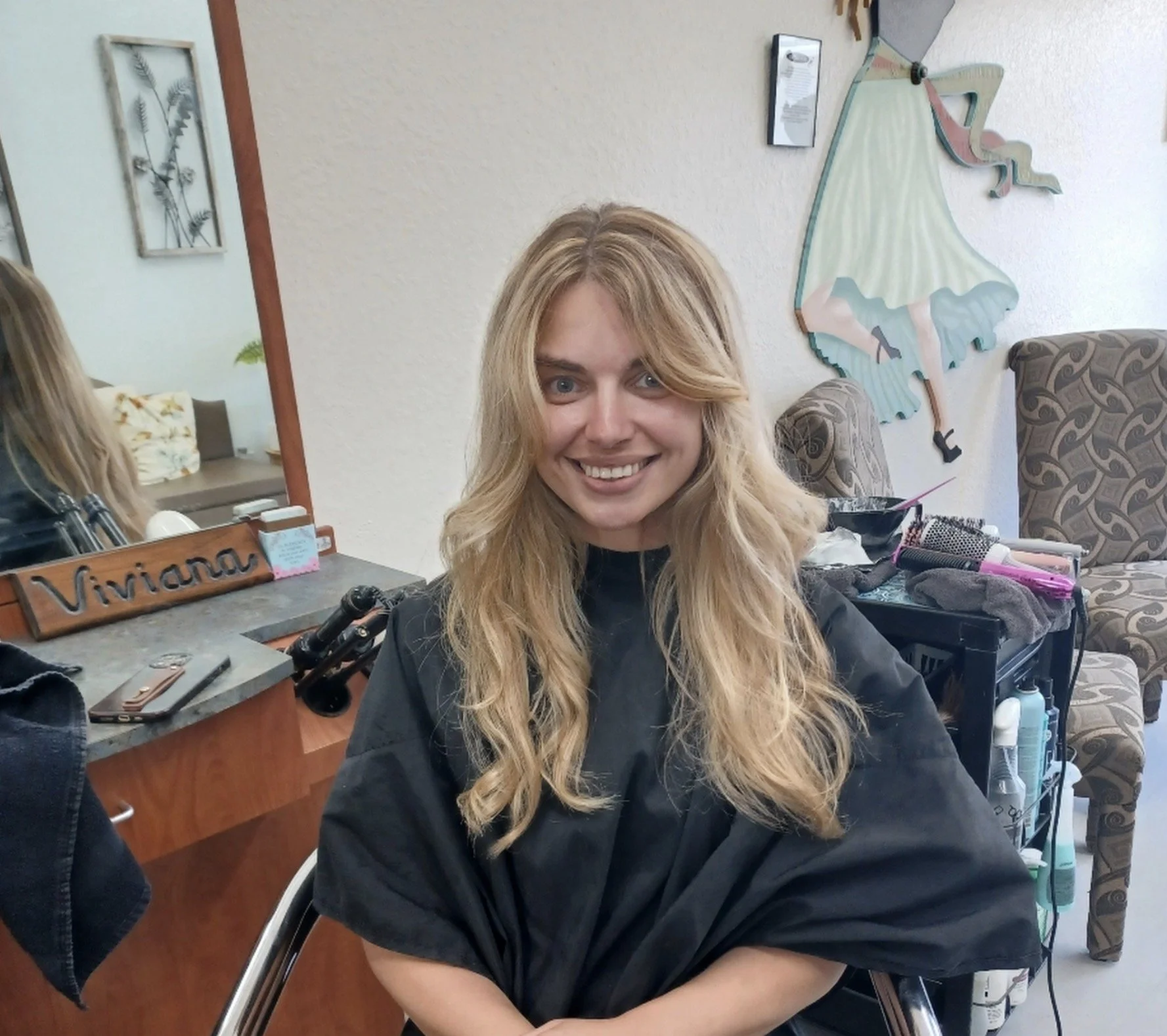 A woman with long blonde hair smiling, sitting in a hair salon chair, with hair styling tools and products on a nearby table. A mirror, framed pictures, and wall art are visible in the background.