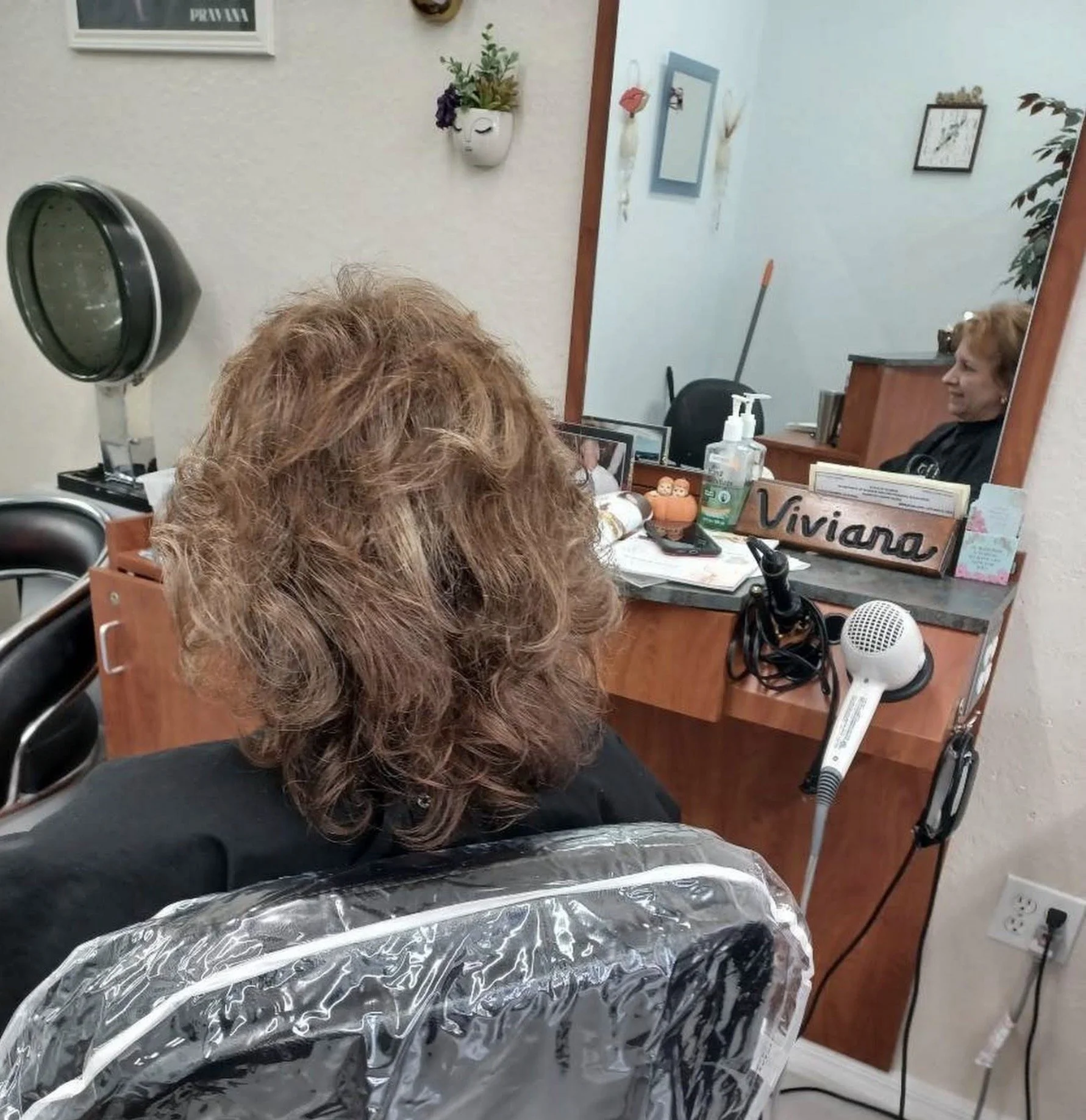 A woman with short curly hair sits at a salon table with her back to the camera, covered with a black cape lined with aluminum foil, receiving a hair treatment. The salon mirror reflects her smiling face, and various hair and beauty products are arra