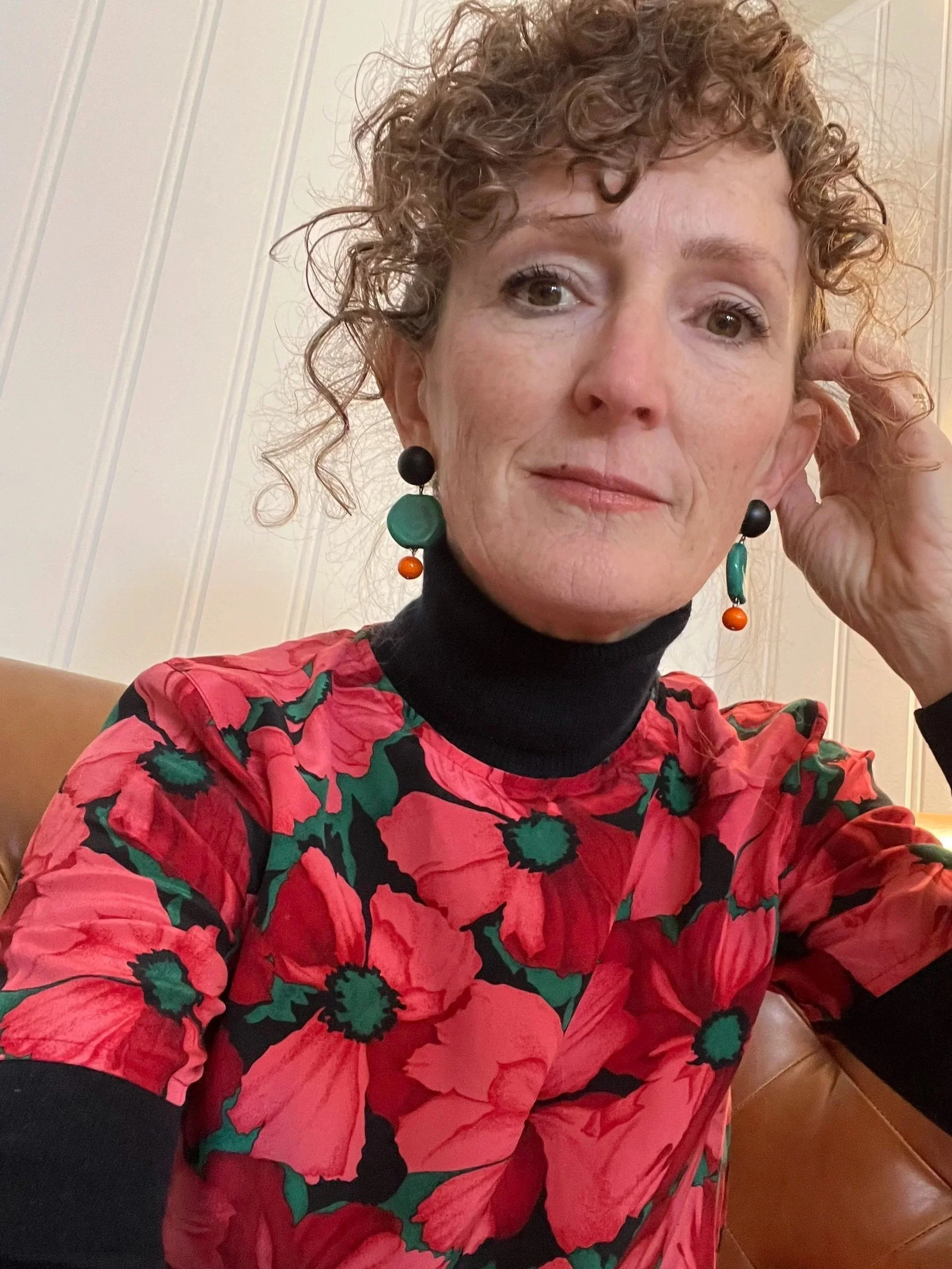 A woman with curly red hair wearing black earrings with green and orange accents, a red floral blouse, and a black turtleneck underneath, sitting on a brown leather chair in front of a white paneled wall.