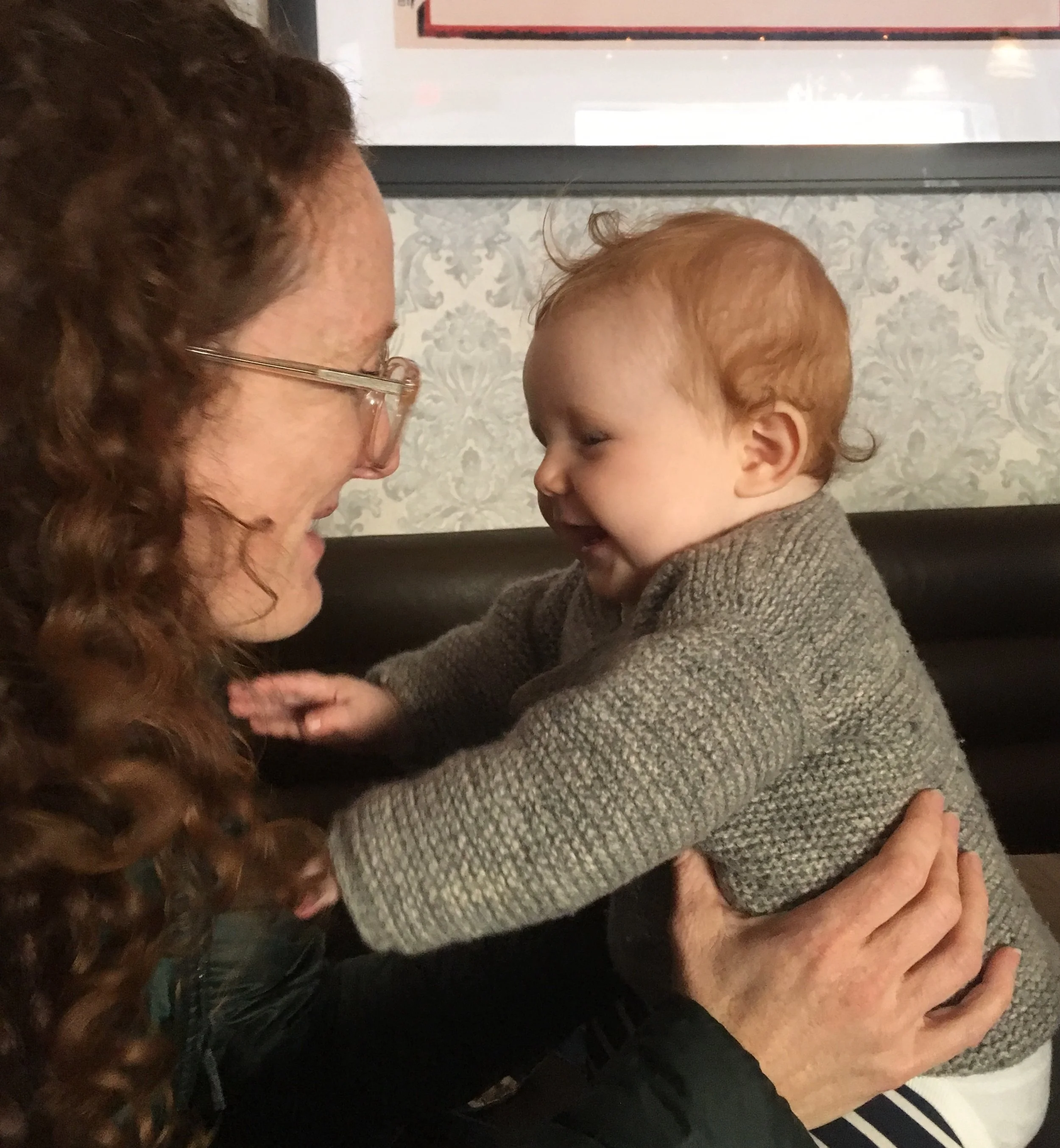 A close-up image of an elderly woman and a young child with curly red hair, facing each other and smiling. The woman wears glasses and a dark jacket, and the child is wearing a gray knitted sweater. The background shows a patterned wall and a television screen.