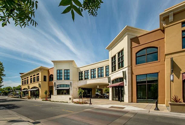 Exterior view of a modern shopping center with storefronts, sidewalk, and street in a suburban area under a blue sky.
