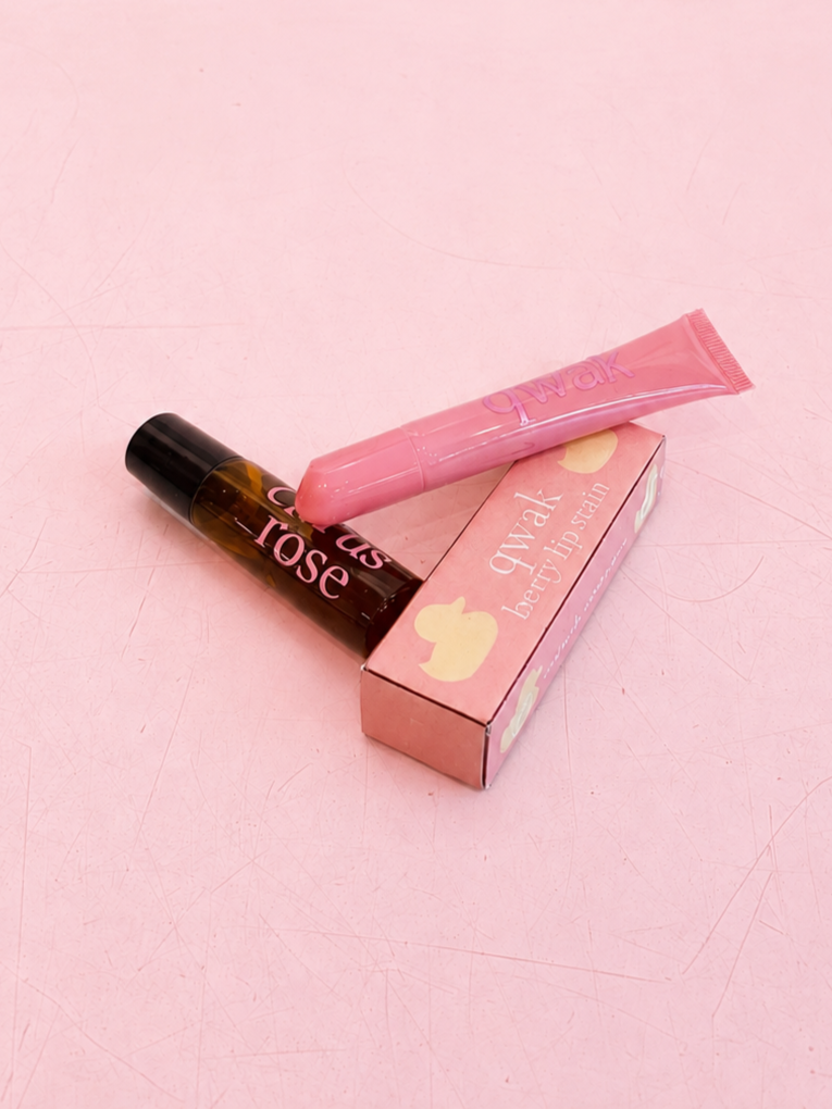 A pink tube of lip gloss and a small bottle of rose scented lip balm placed on a pink box with a yellow duck illustration. The items are on a pink surface.
