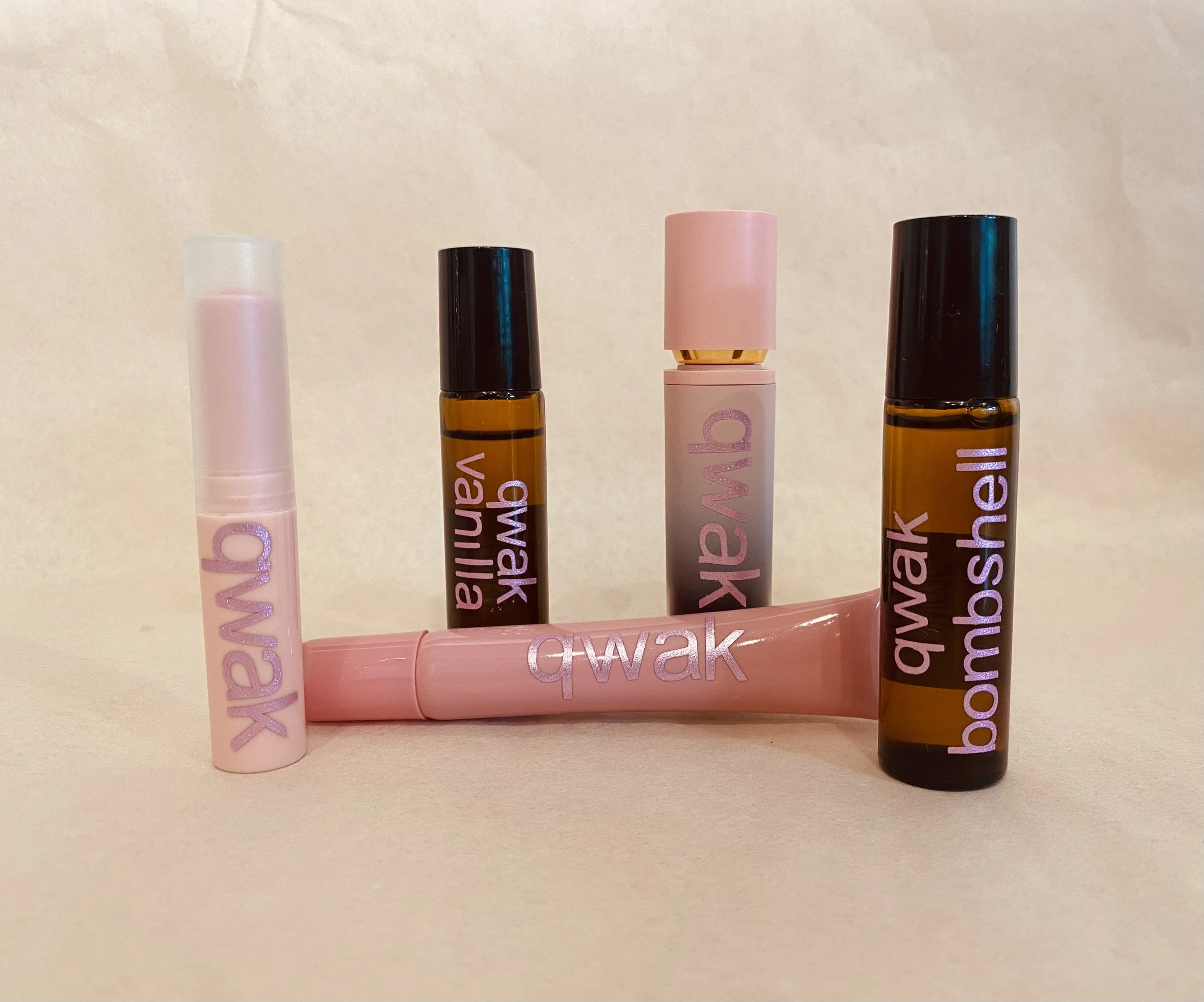 Six skincare or cosmetic tubes arranged in a row on a pale beige background, with a small pink bottle lying horizontally below the row.