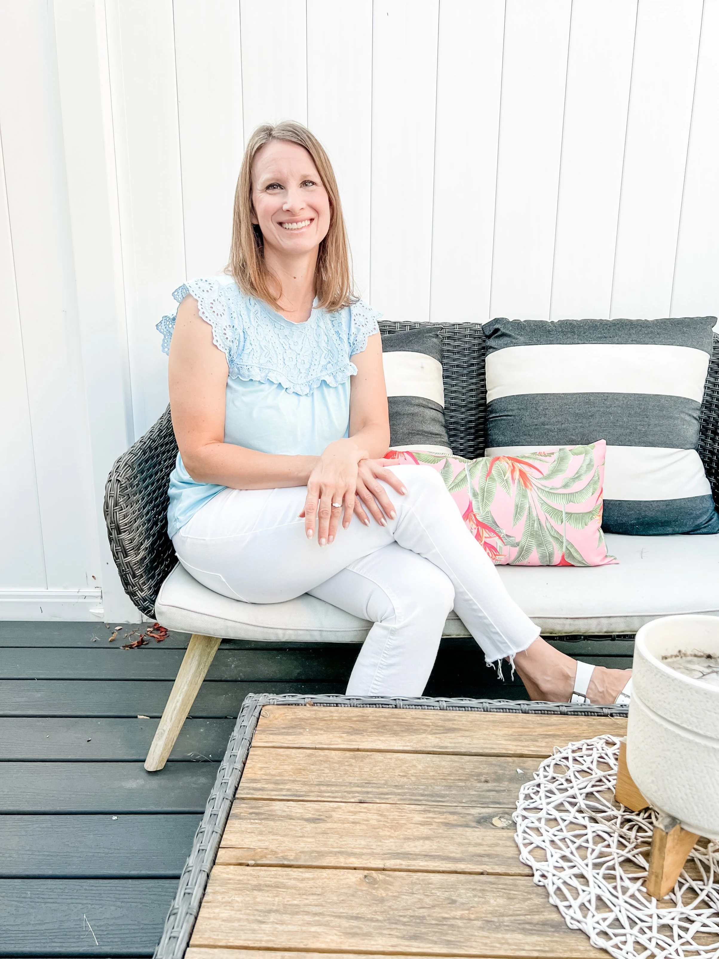 A woman sitting on a cushioned outdoor wicker chair, dressed in a light blue top and white pants, smiling, with colorful pillows behind her on a patio with white paneled walls and a dark wood deck, a wooden table in front, and a decorative white wicker vase on a side table.