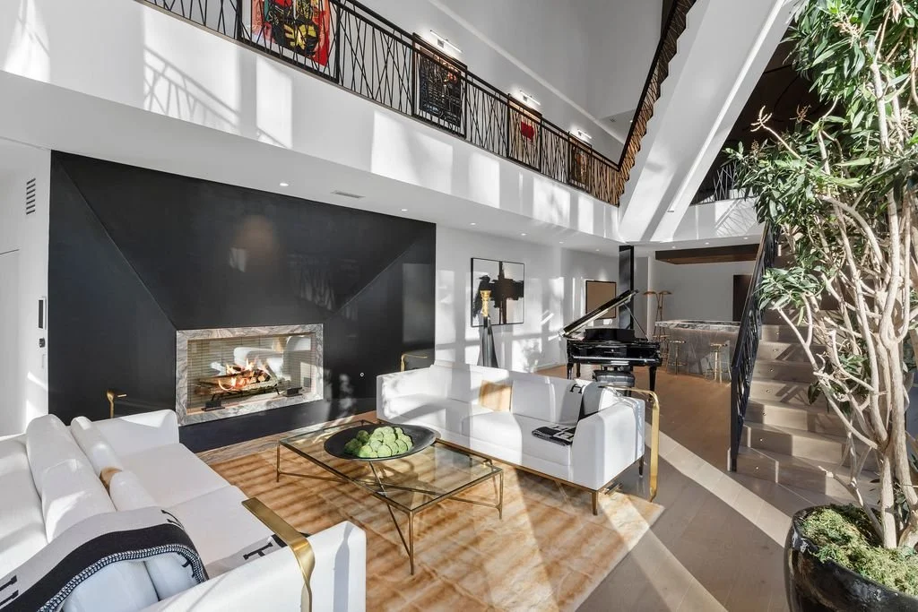 Modern living room with white sofas, a glass coffee table, a black fireplace, a grand piano, and a staircase with wooden steps and black railing, decorated with artwork on the walls and greenery nearby.