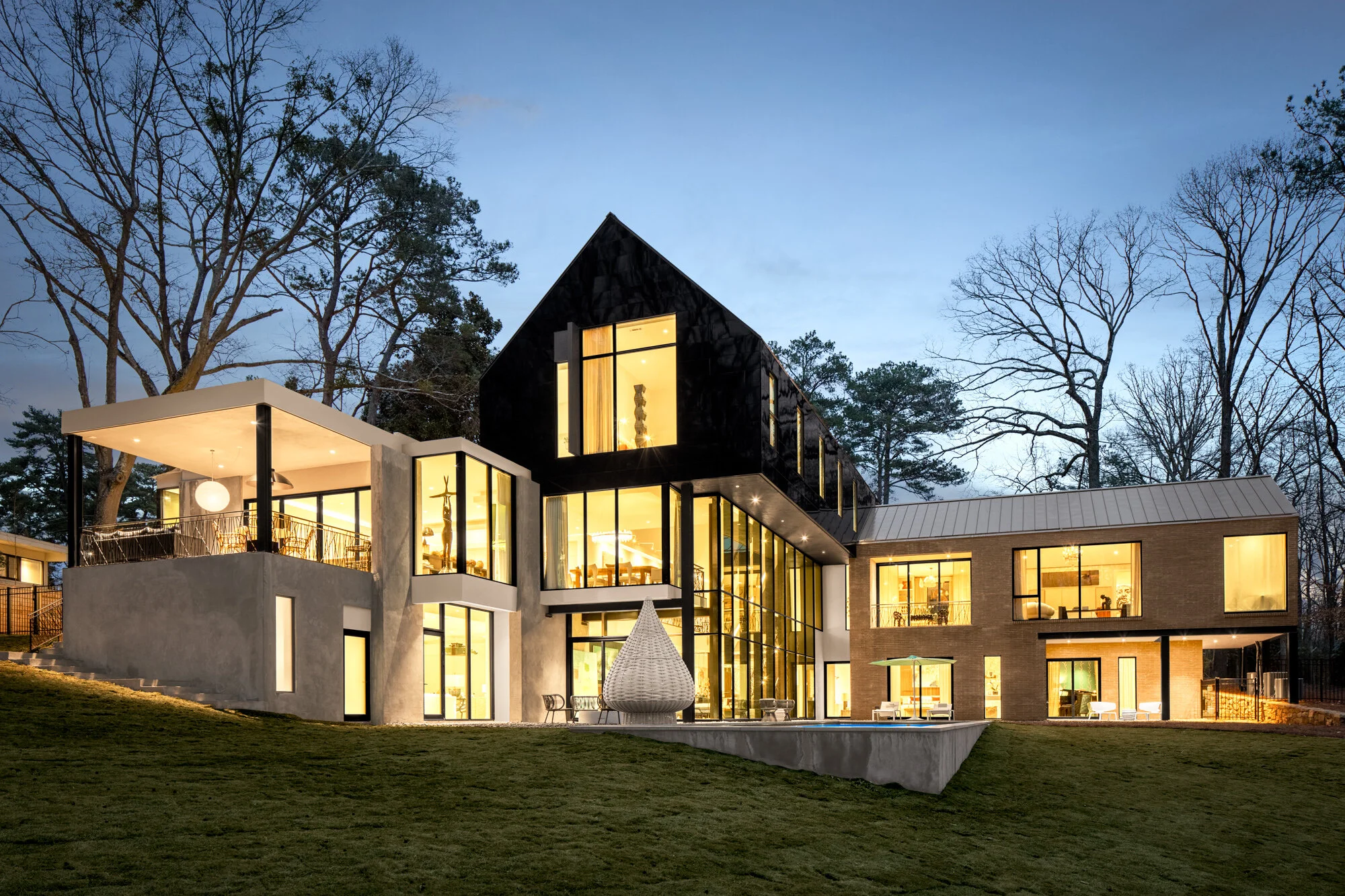 Modern multi-story house with large windows and outdoor patio area illuminated at dusk, surrounded by trees.