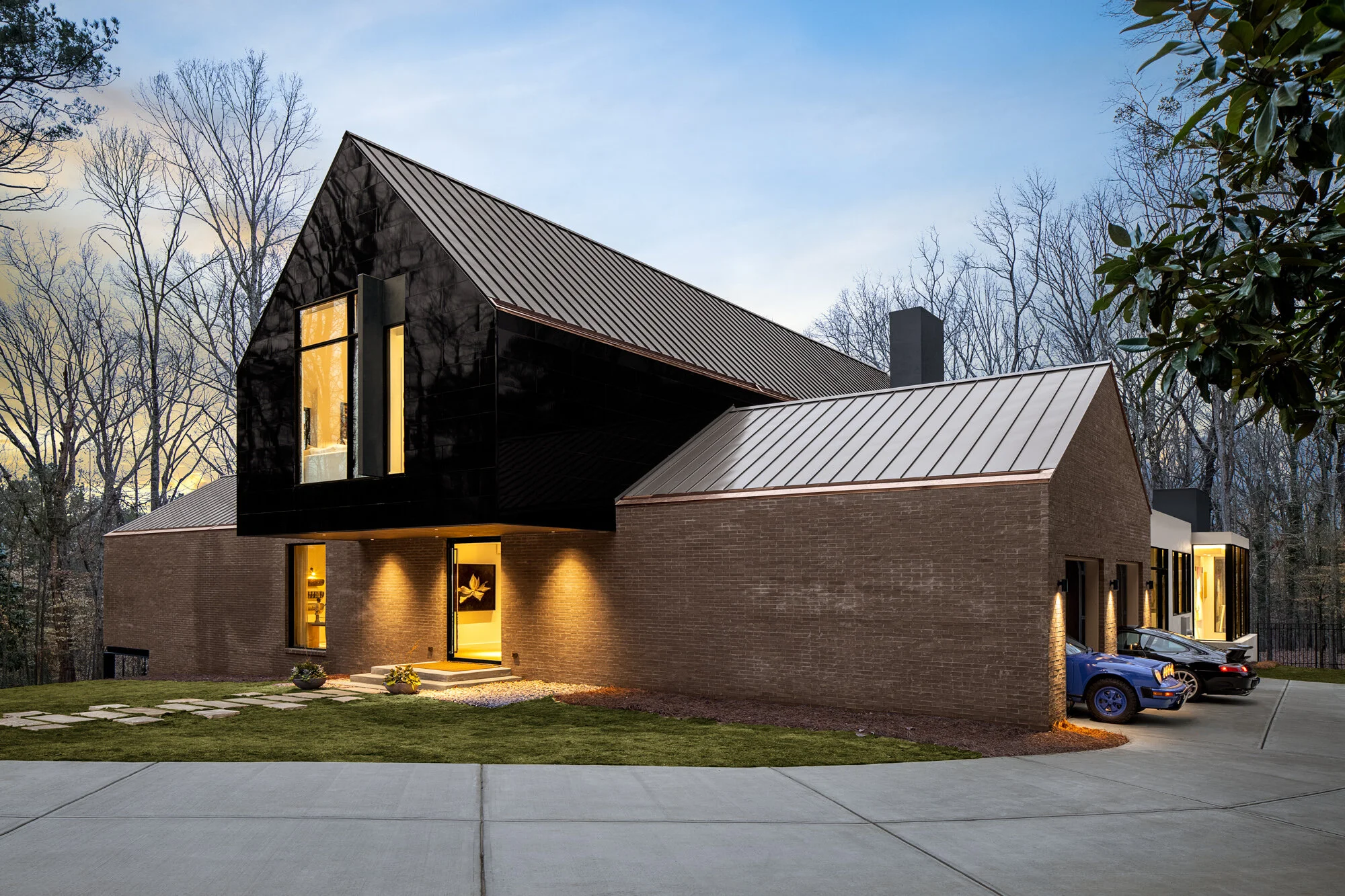 Modern house with brick walls, black window frames, and metal roofs, illuminated from inside during dusk, surrounded by trees and a well-maintained lawn, with a driveway and parked cars in the background.