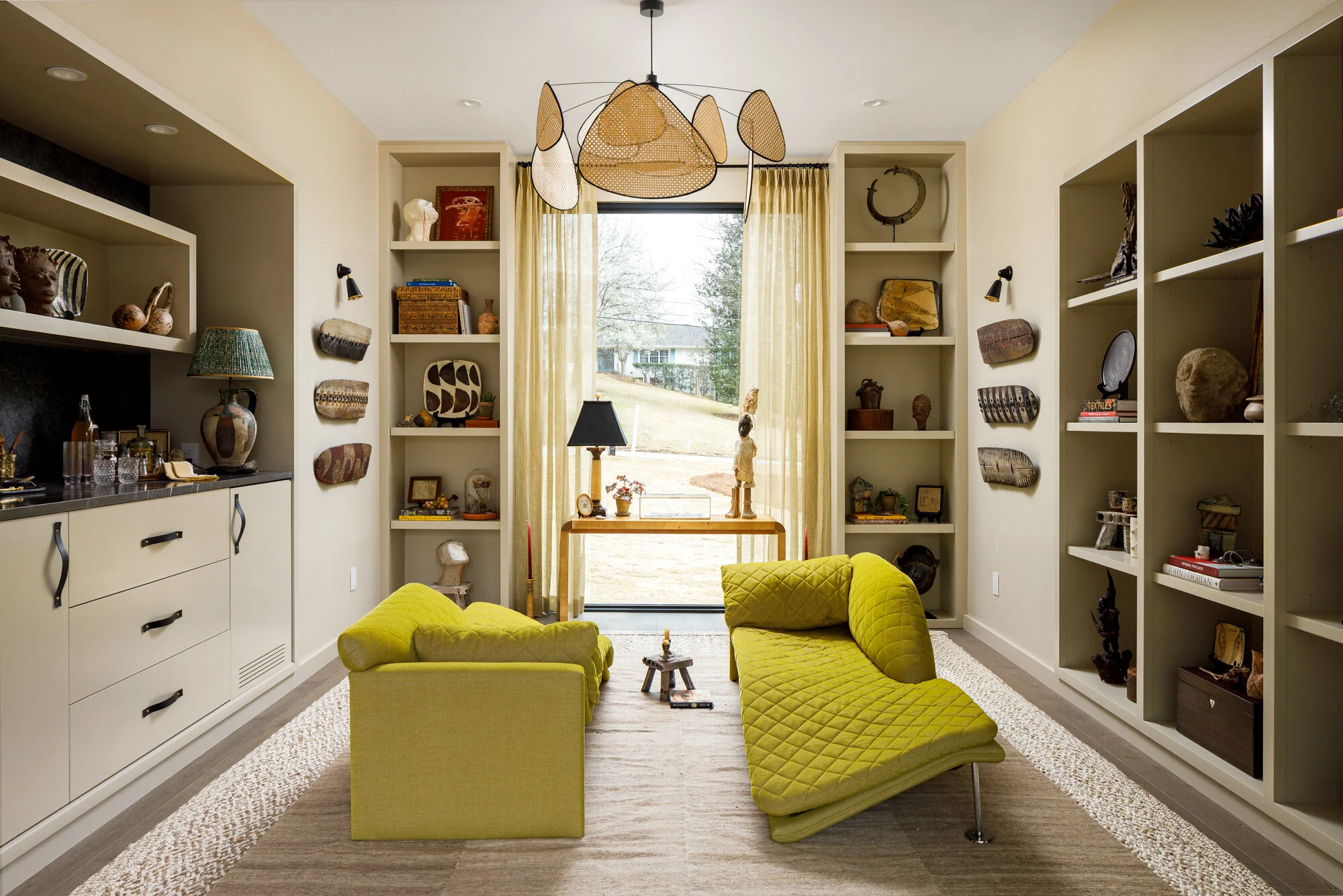 Living room with yellow quilted chairs, creambookshelf with art and decor, large window with yellow curtains, modern chandelier, and eclectic decor pieces.