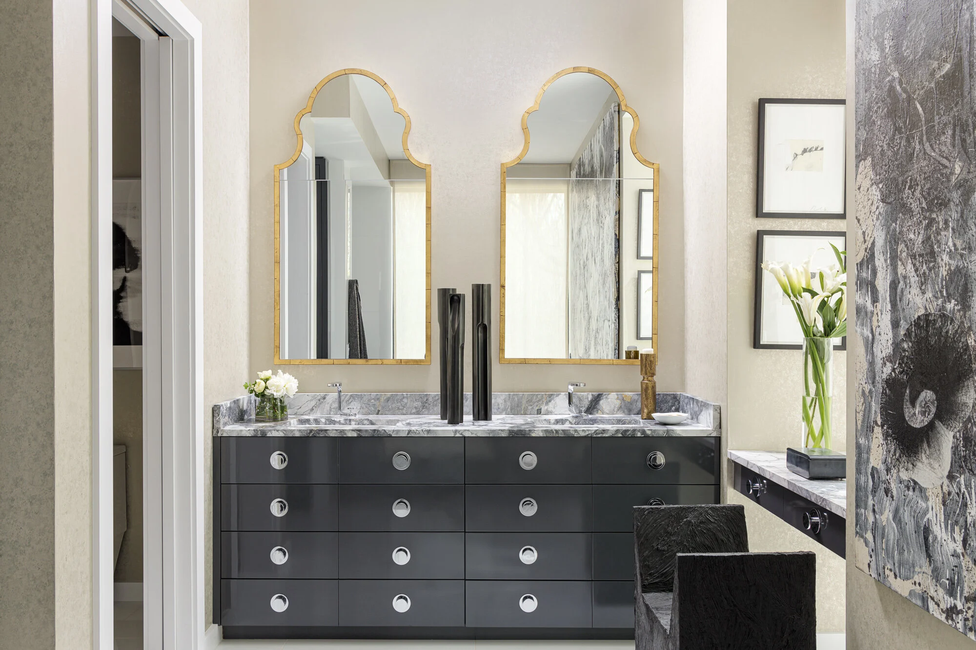A modern bathroom vanity with two large mirrors accented with gold trim, a marble countertop, black cabinetry with circular silver handles, and decorative vases and plants.