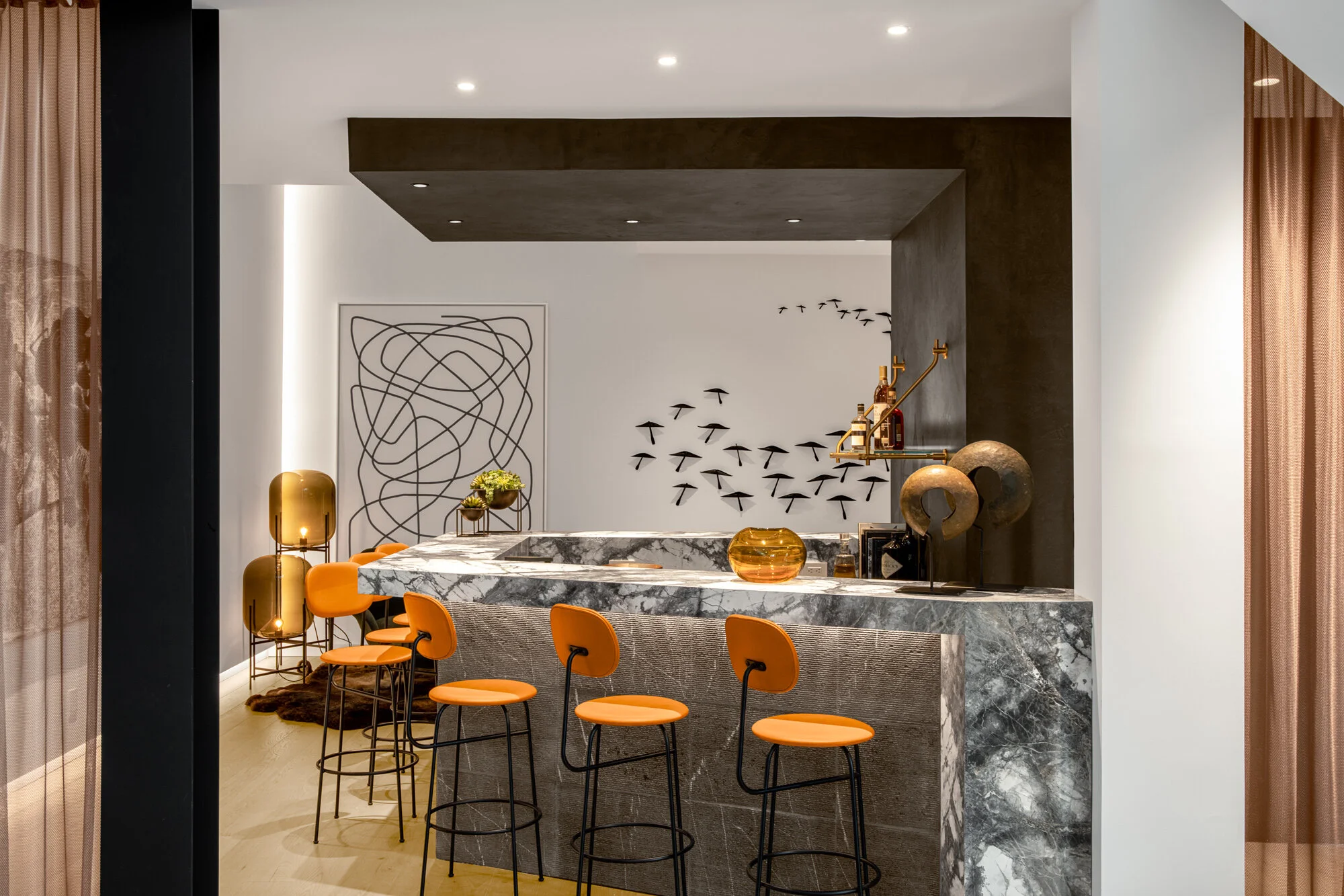 Modern bar area with a marble counter, orange bar stools, and decorative wall art including a tree and birds.