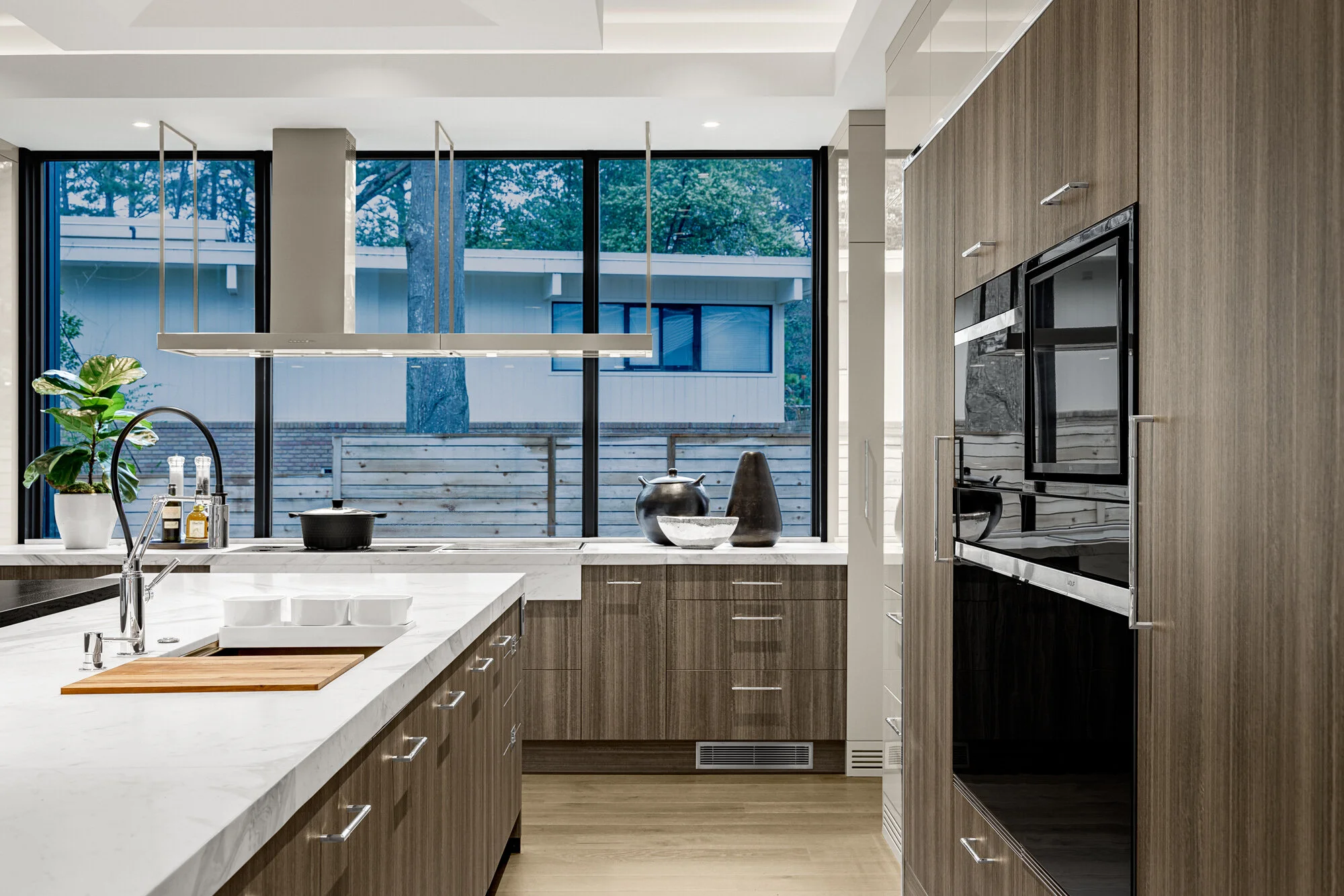 Modern kitchen with large windows, white marble countertops, wooden cabinets, and black appliances. Decor includes potted plant, bowls, and kitchen utensils.