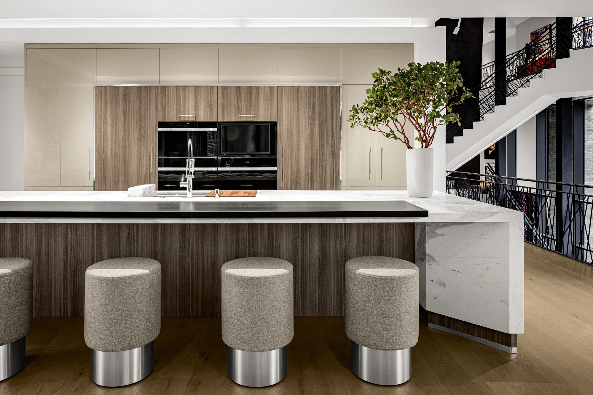 Modern kitchen with beige and wood cabinets, a large kitchen island with a white marble top, four beige upholstered bar stools, a potted green plant, and a staircase with black railings in the background.