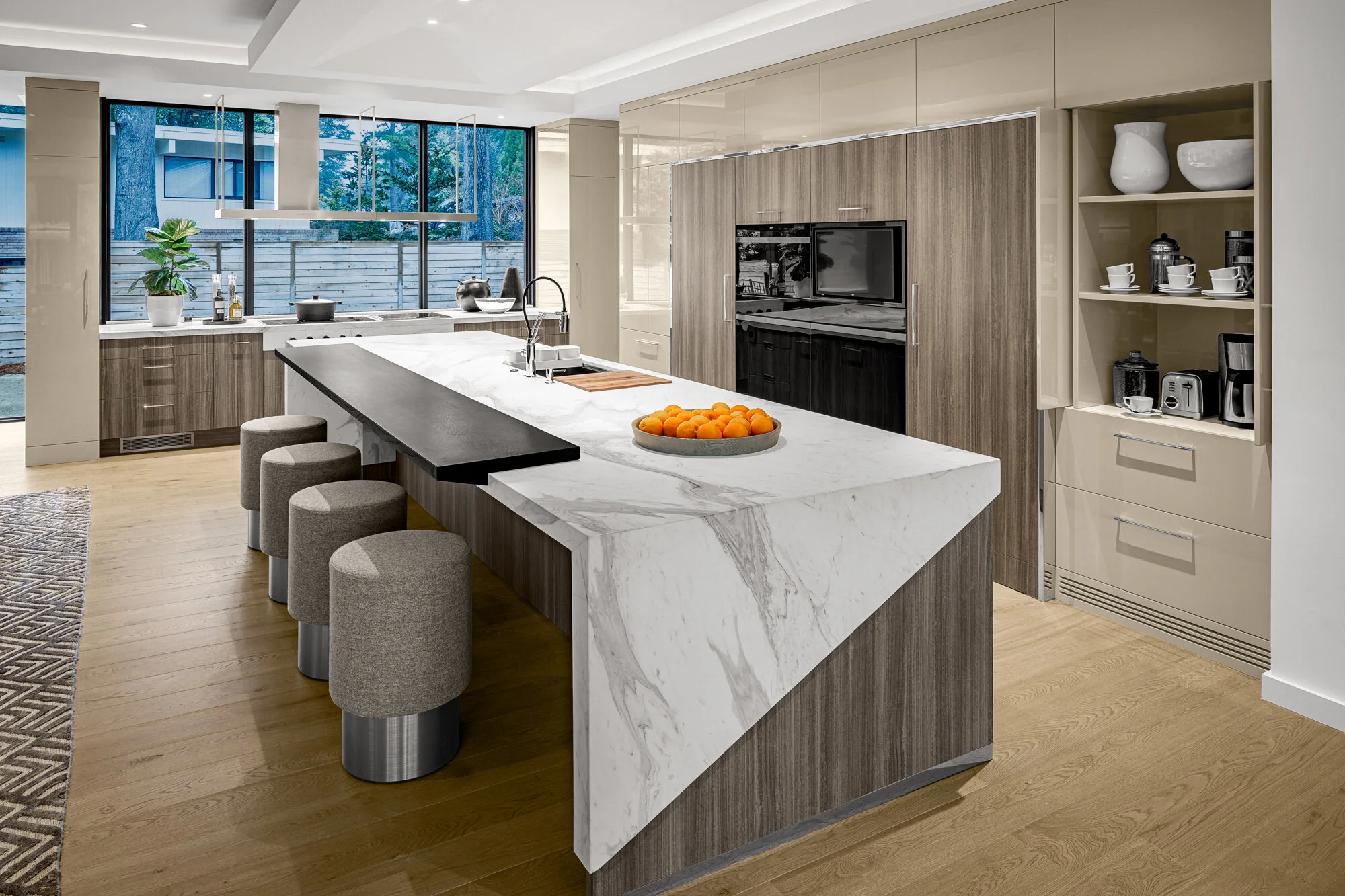 Modern kitchen with a large island with a white marble countertop, surrounded by gray upholstered bar stools. The back wall features floor-to-ceiling cabinets and open shelving with dishes and appliances. Large windows provide natural light and a vie