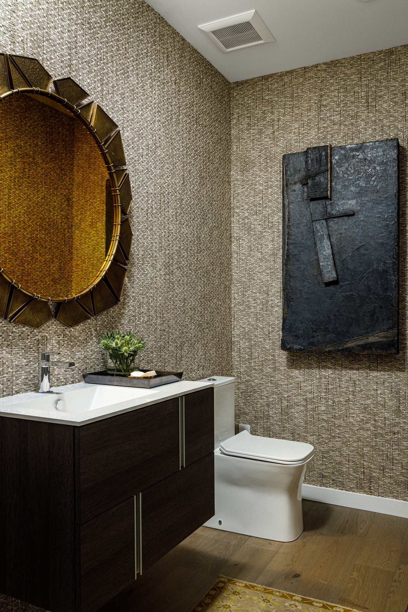 A modern bathroom featuring textured wallpaper, a dark wood vanity with a white sink, a circular mirror with a gold frame, a small plant, a black tray, a white toilet, a black textured wall art piece, a ceiling vent, and a small yellow rug on wooden 