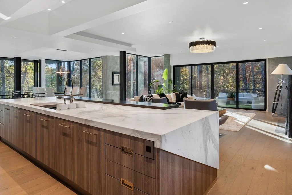 Modern open-concept living space with a kitchen island, dining area with chairs, and a living room with sofas, large windows showing outdoor trees, and contemporary decor.