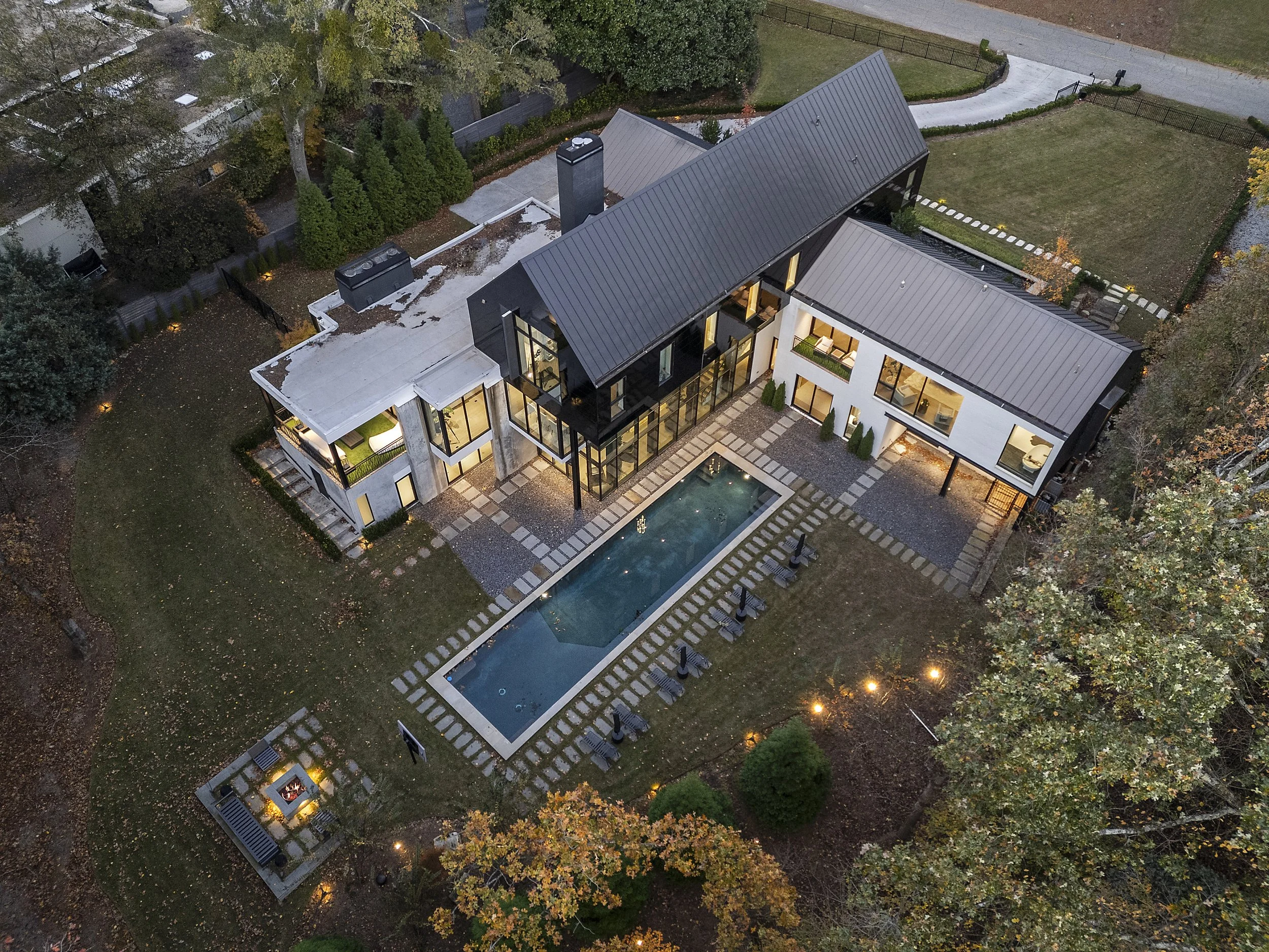 Aerial view of a modern house with a swimming pool, surrounded by a backyard with trees, grass, and a pathway, with late evening lighting.