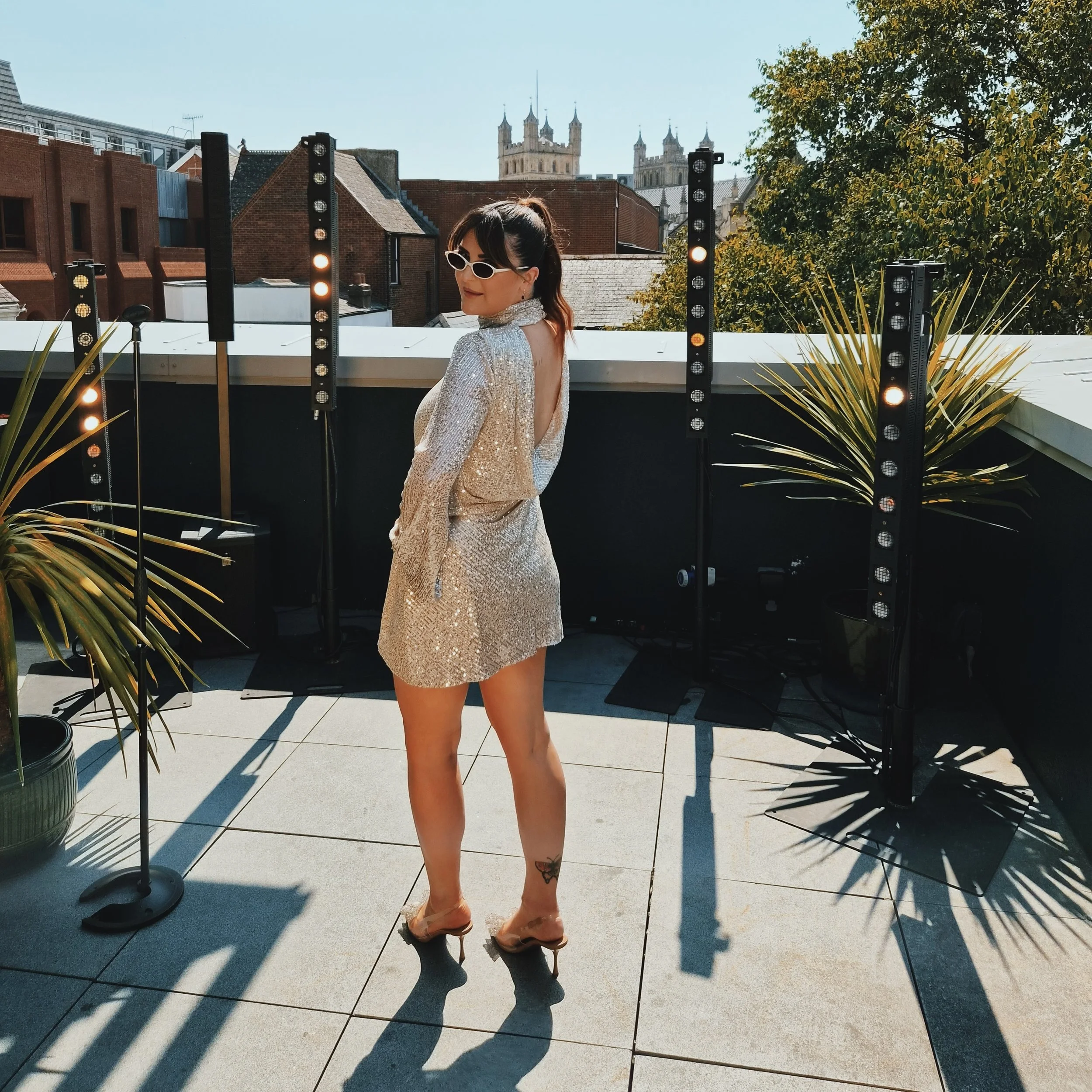A woman in a sparkly dress and sunglasses standing outdoors on a sunny day, with a city skyline and trees in the background.