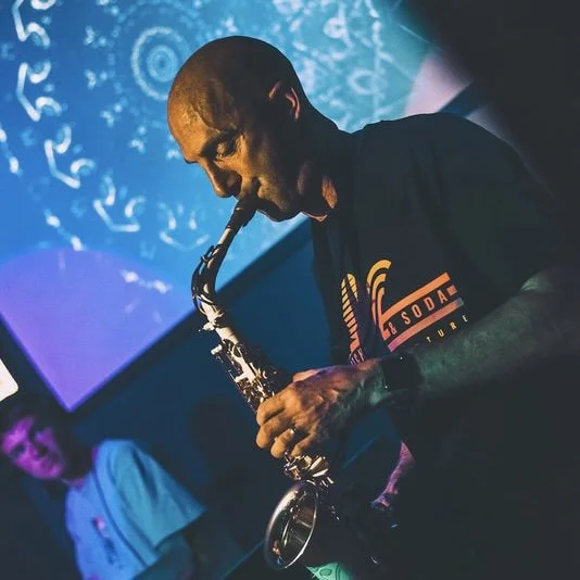 A man playing a saxophone during a live music performance with colorful lighting and an audience member in the background.