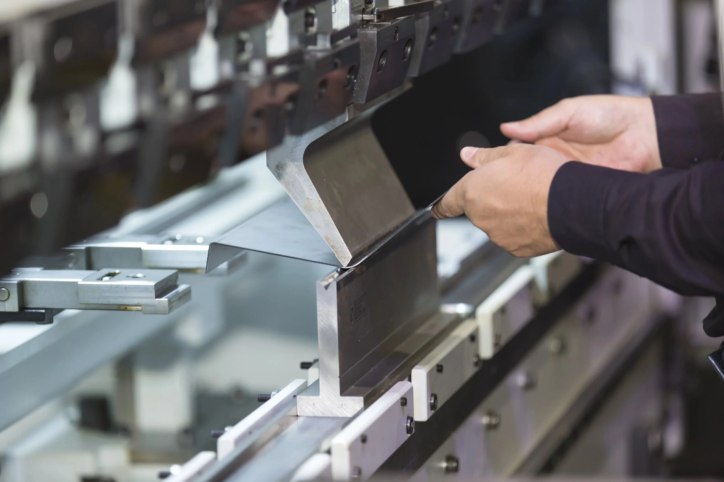 Close-up of a person's hands holding a metal sheet near an industrial machine with multiple metal components.