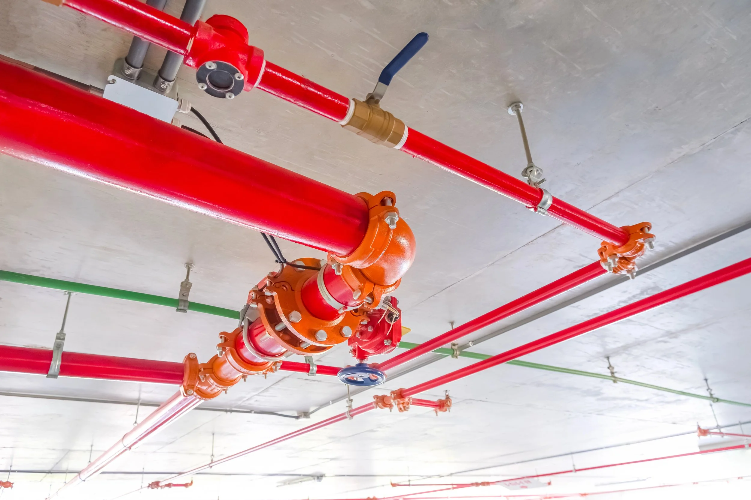 Red and orange pipes and valves attached to a concrete ceiling, with a blue handle and green pipes in the background.
