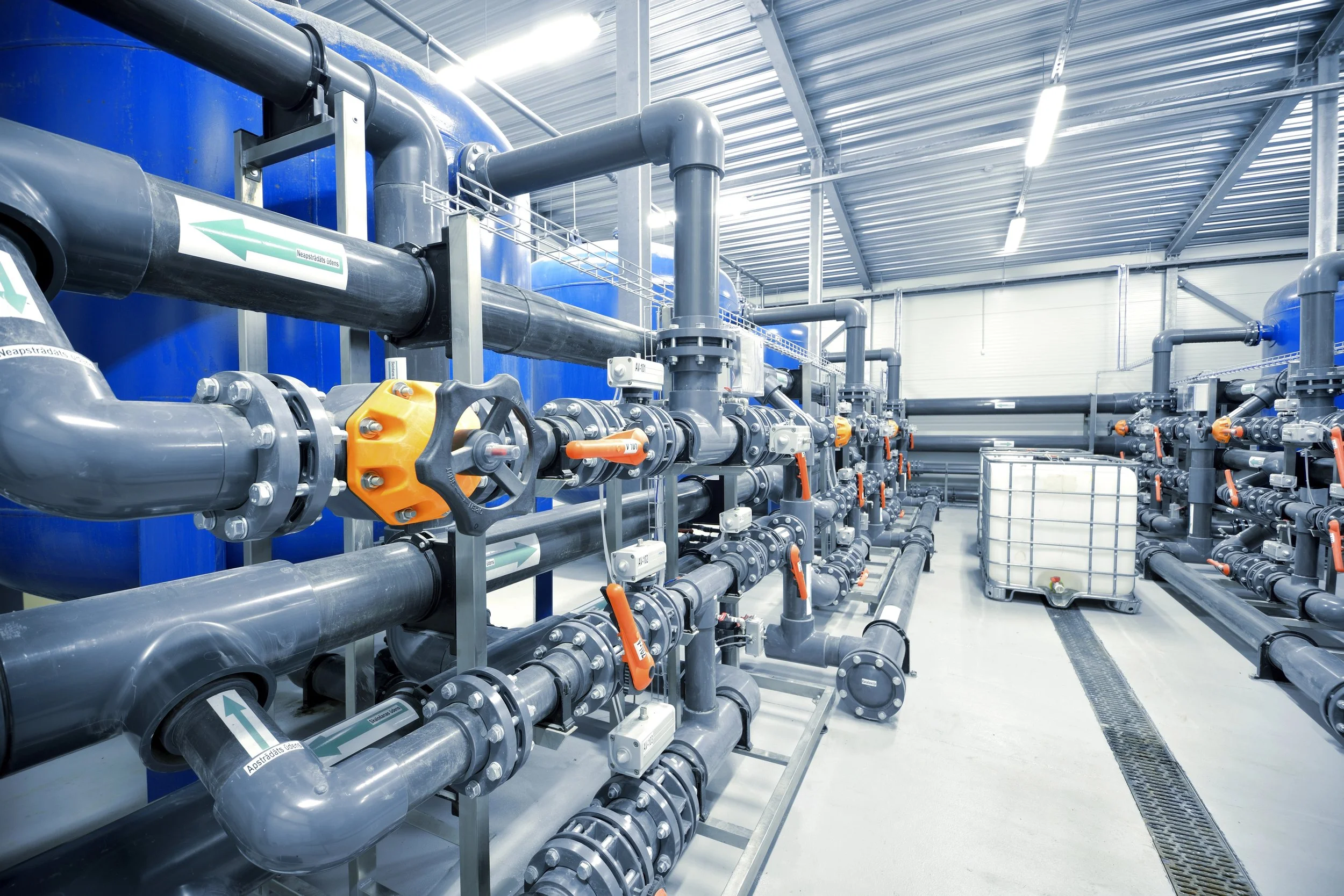 Industrial facility with large gray pipes, valves, and blue tanks, inside a metal building with a corrugated metal roof.