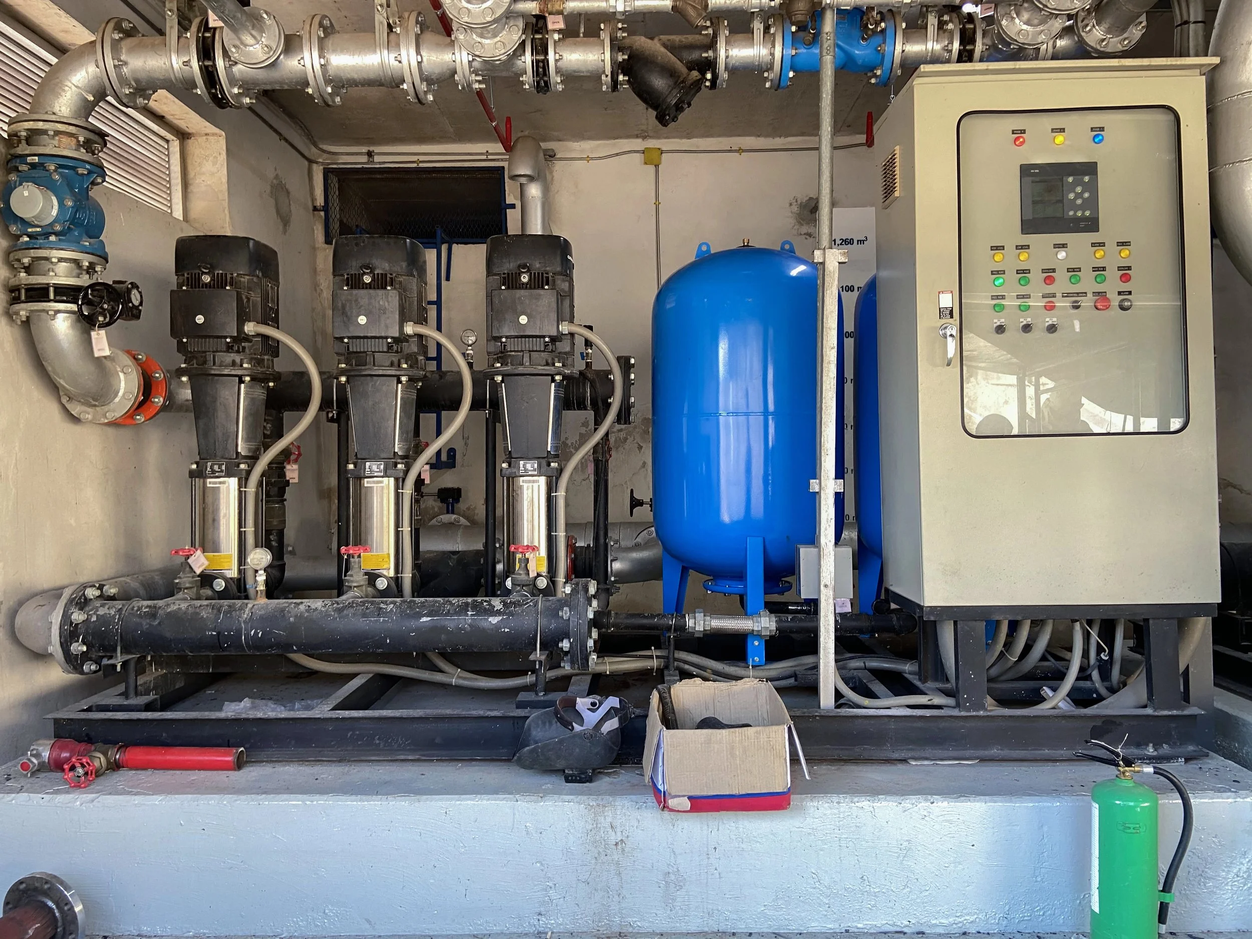 A mechanical room with pipes, pumps, tanks, and a control panel. Contains three black pumps, a blue tank, various pipes, valves, and electrical wiring.