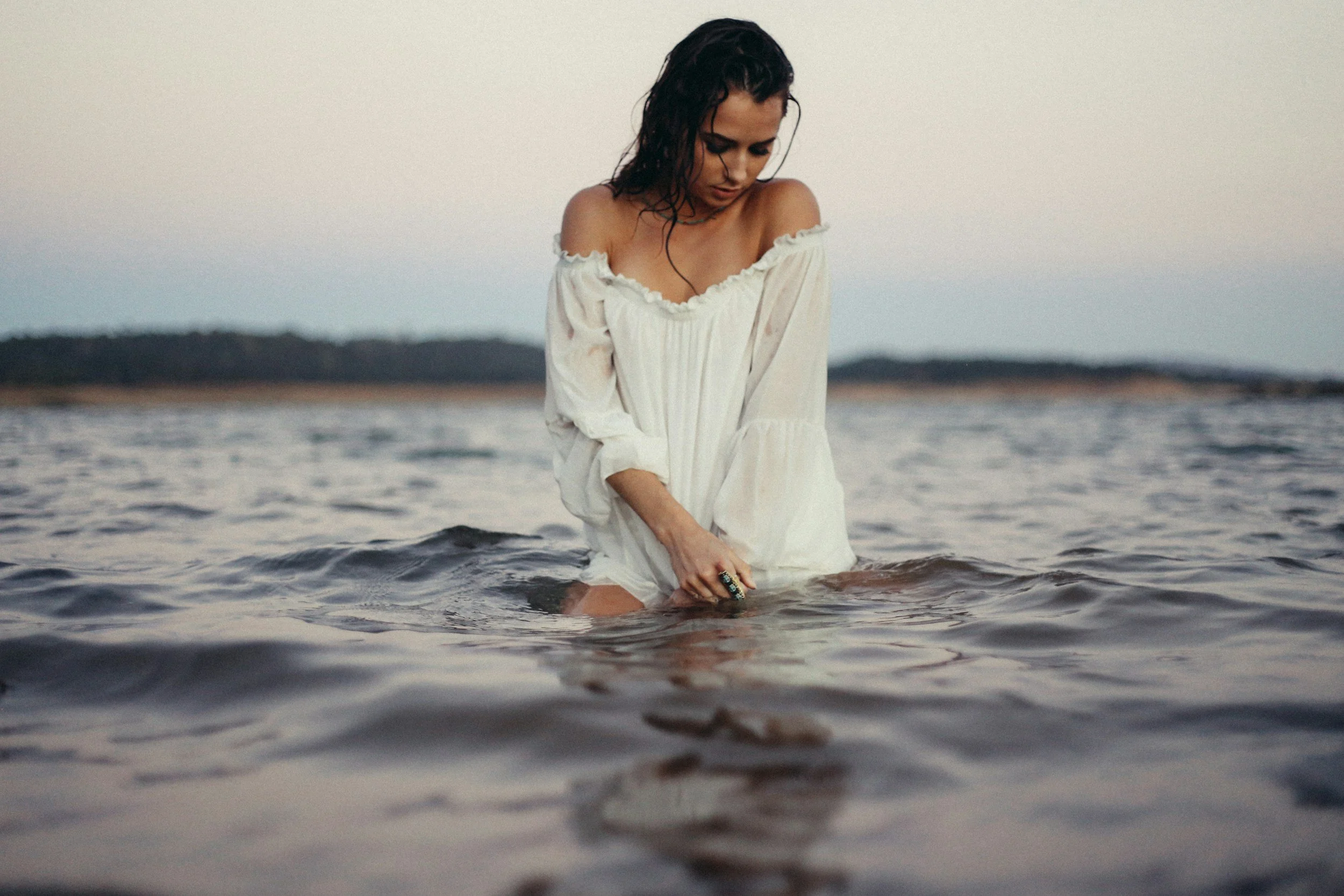 Woman wearing a white off-shoulder dress standing in water with distant shoreline in the background.
