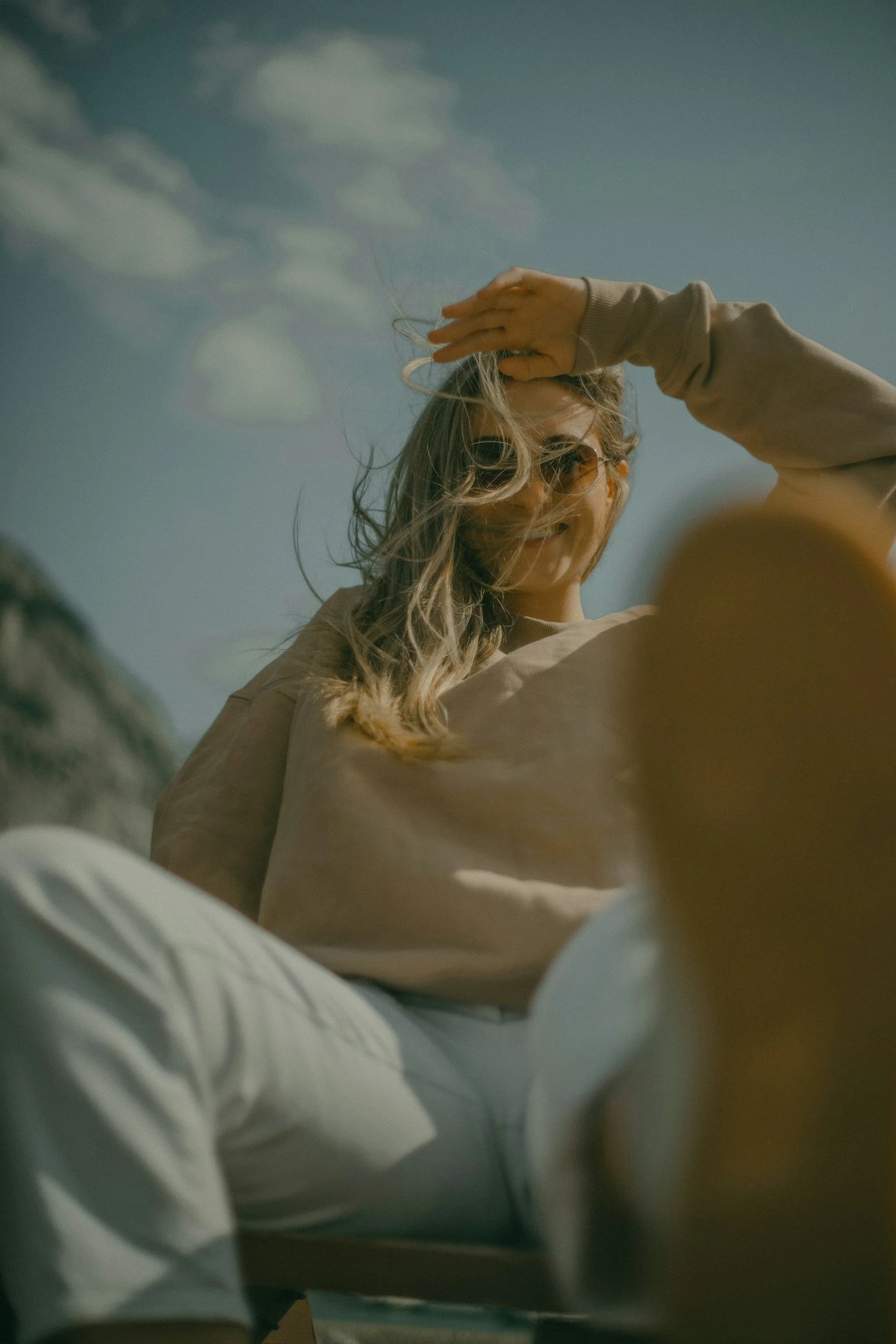 A woman with blonde hair and sunglasses takes a selfie outdoors against a blue sky with clouds. She is wearing a light-colored long-sleeve shirt and appears to be smiling, with her hand on her head.