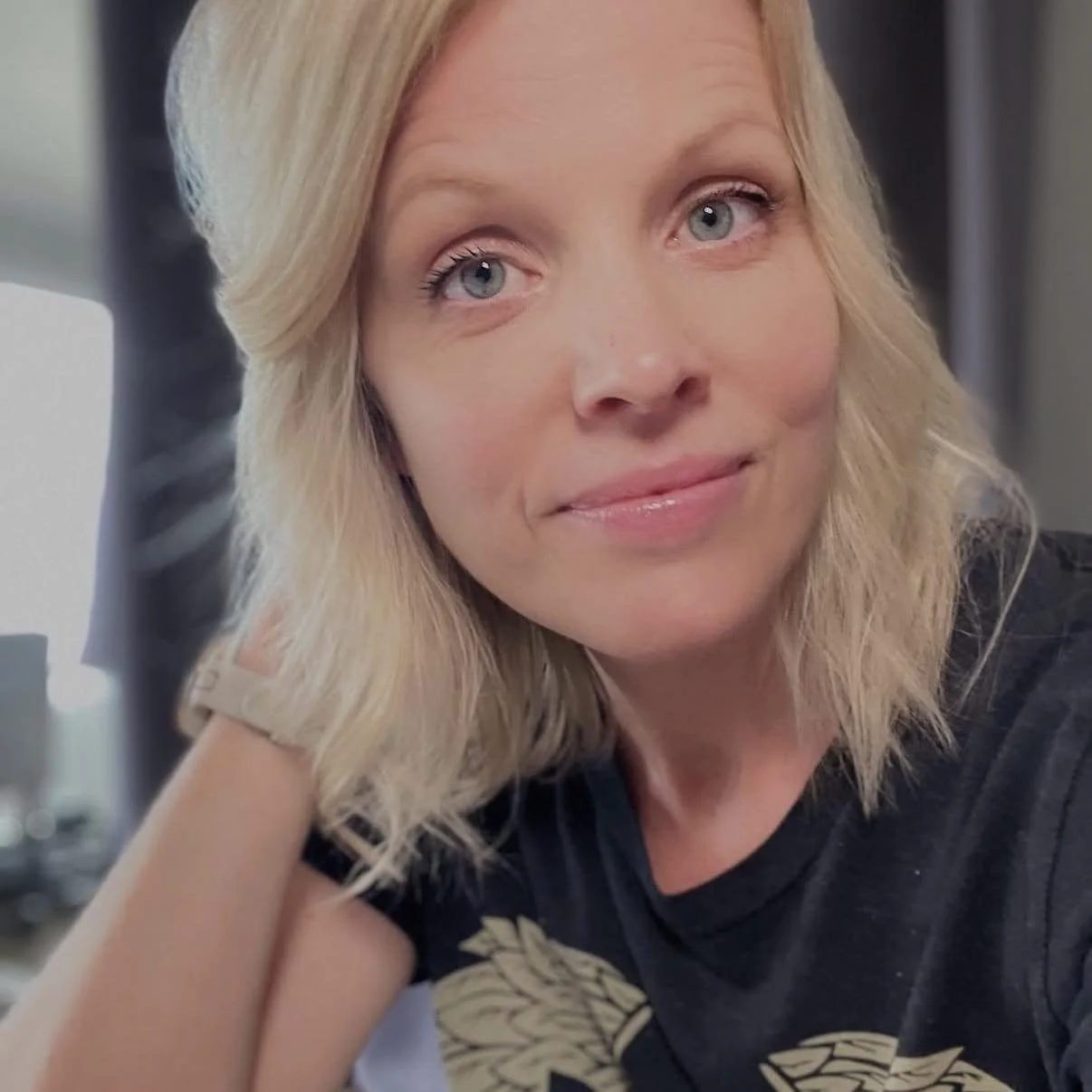 A blonde woman with blue eyes, wearing a black t-shirt, smiling softly and resting her head on her hand in a casual indoor setting.
