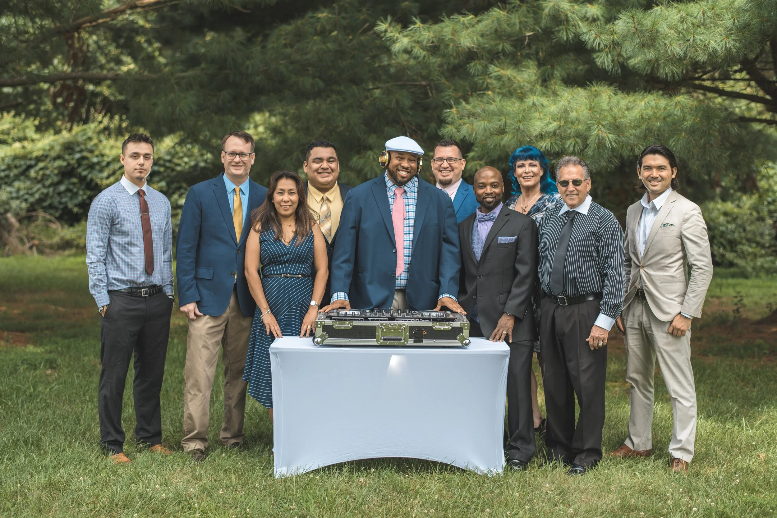 A diverse group of eleven people, including men and women, standing outdoors in front of green trees, dressed in formal to semi-formal attire, with DJ equipment on a table in front of them, smiling at the camera.