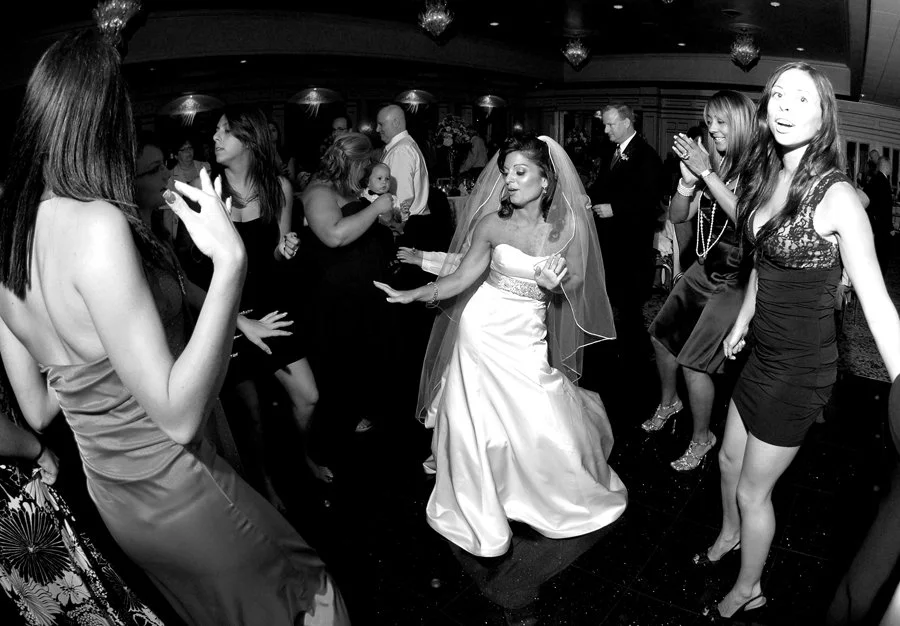 A wedding reception with women dancing and celebrating, including a bride in a white wedding gown and veil. The guests are dressed in formal attire and appear to be enjoying the party.