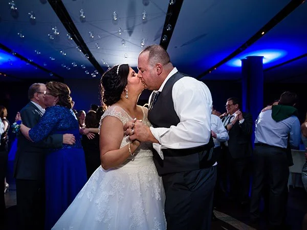 Bride and groom sharing a kiss during their wedding reception, surrounded by guests in formal attire, in a decorated indoor venue.