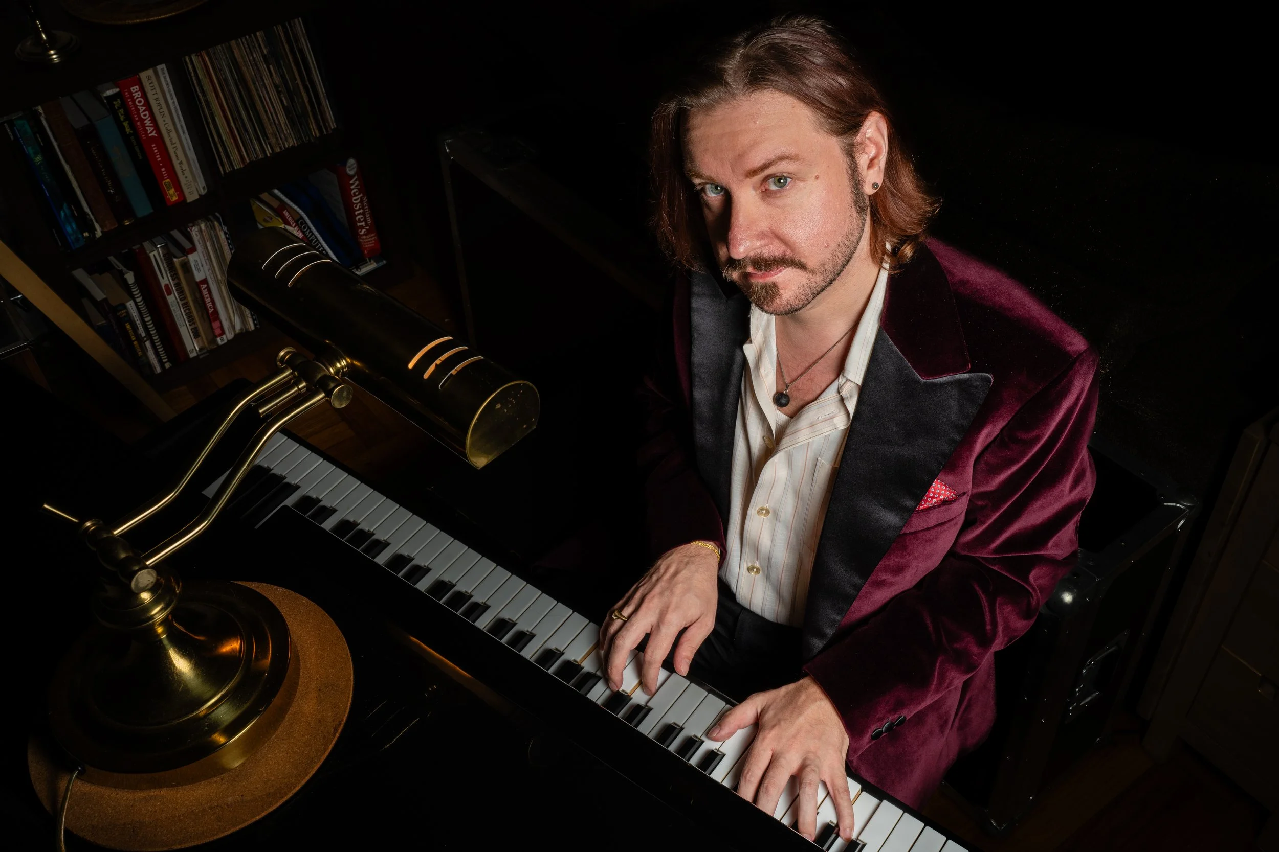 A man with long brown hair and a beard, wearing a maroon velvet blazer and playing a grand piano.