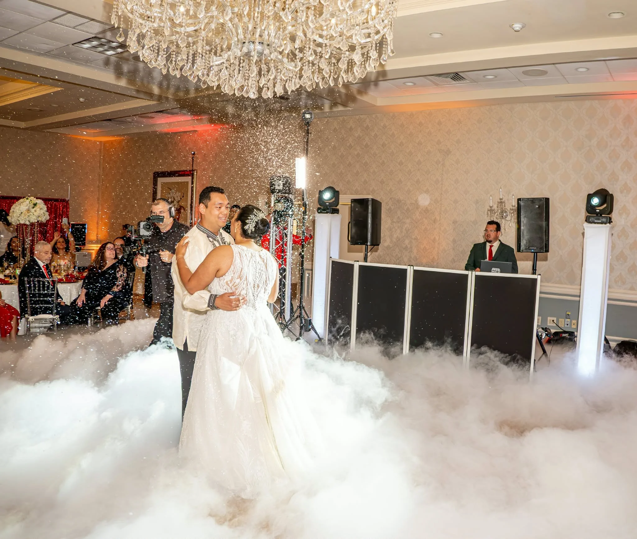 A couple of newly married individuals sharing a dance at their wedding reception, surrounded by guests and celebration decor, with a fog machine creating a cloud on the dance floor.