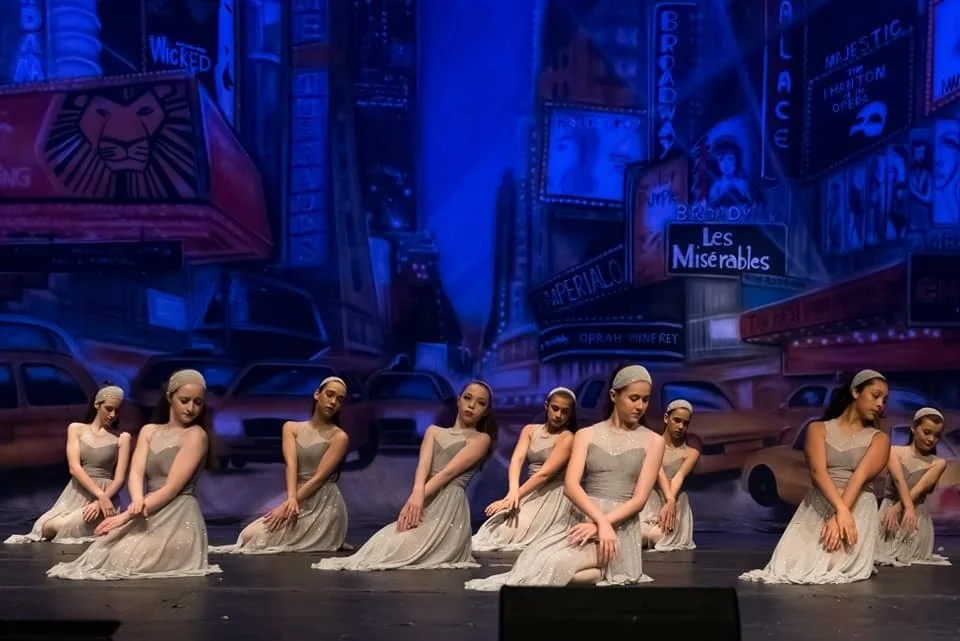 Group of young female dancers in light gray dresses and headbands performing a ballet on stage with a colorful cityscape backdrop featuring billboards, taxis, and Broadway signs.