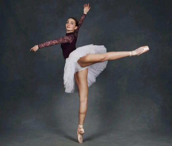 A ballerina in a pose with one leg raised and arms extended, wearing a white tutu, a long-sleeved lace top, and ballet slippers, standing on a gray background.